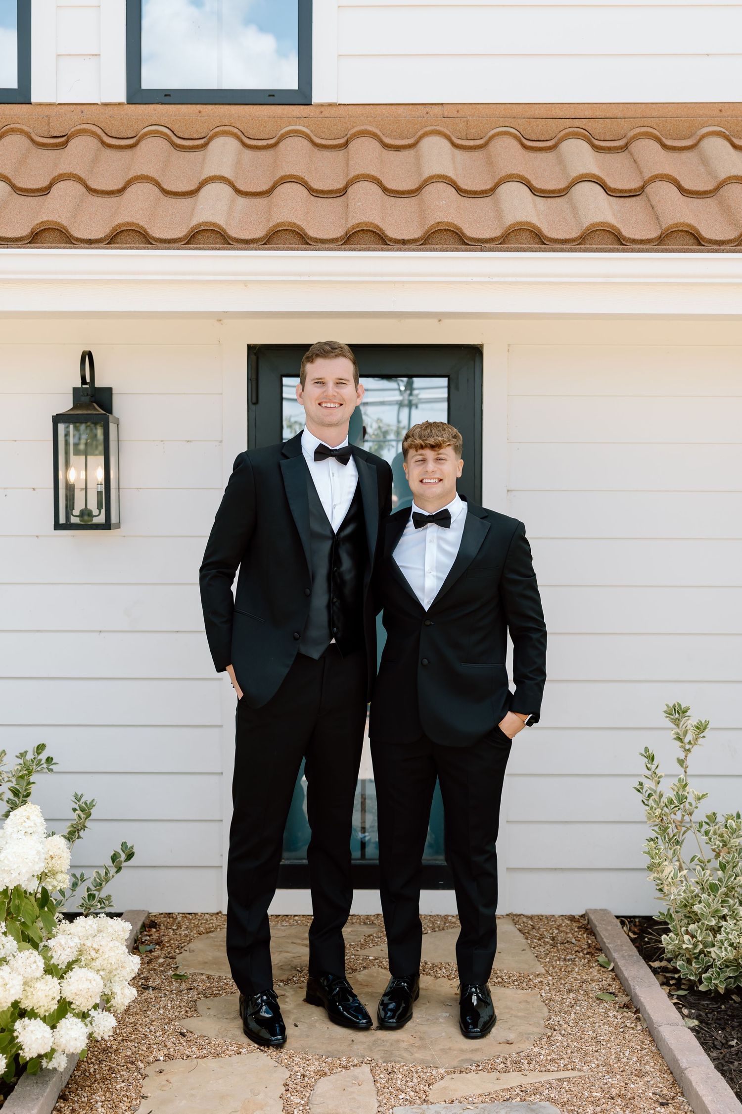 Two young men in black tuxedos with bow ties pose smiling outside a white home with terracotta roof tiles.