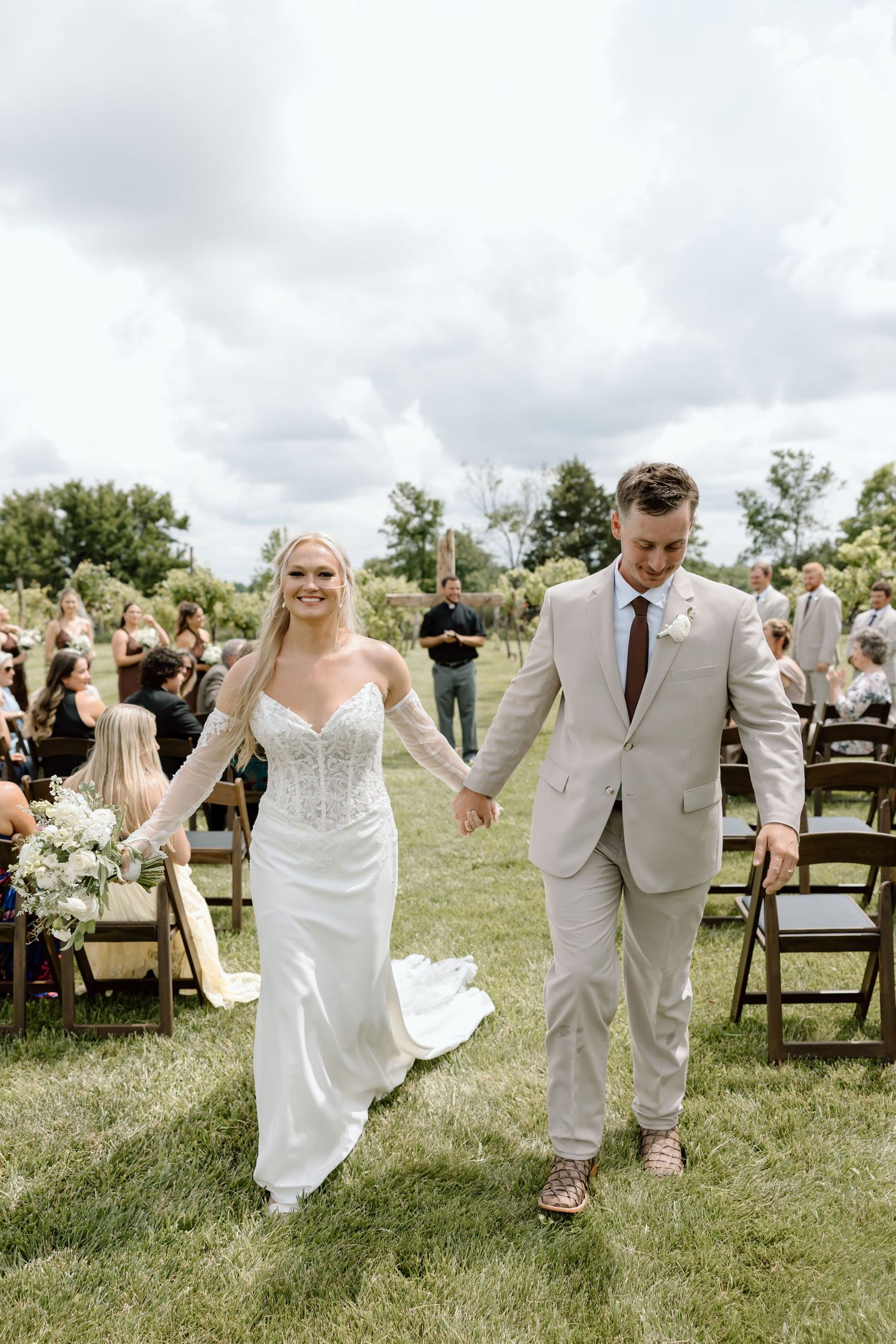 Bride and groom walk hand-in-hand down outdoor wedding aisle, bride in lace gown, groom in tan suit.