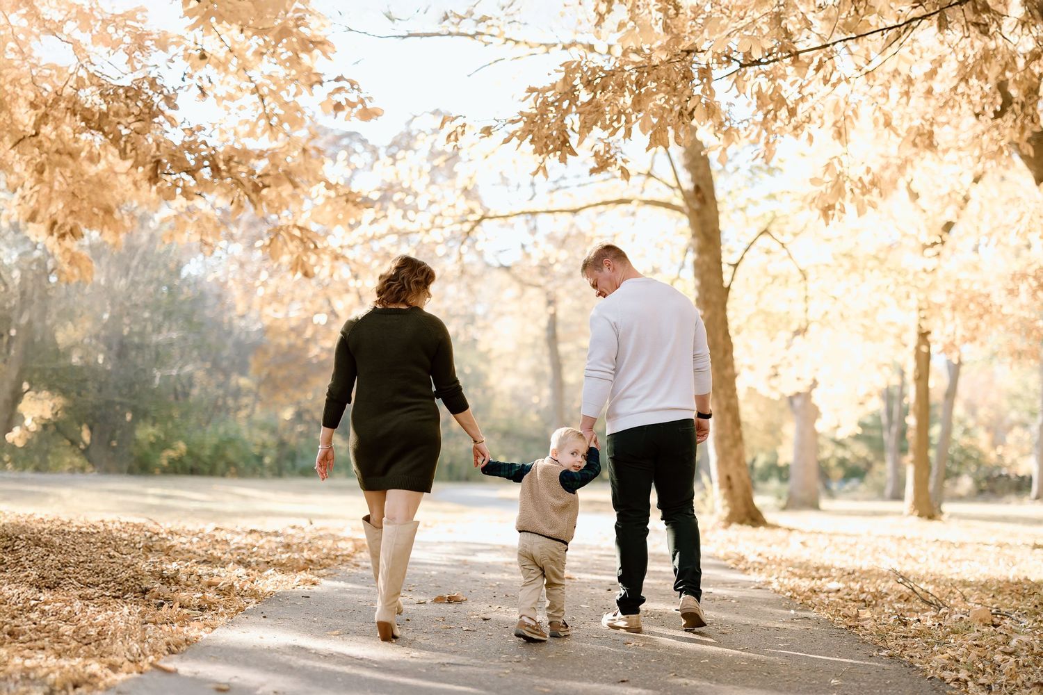 A small child walks between two adults on a tree-lined autumn path with fallen leaves.