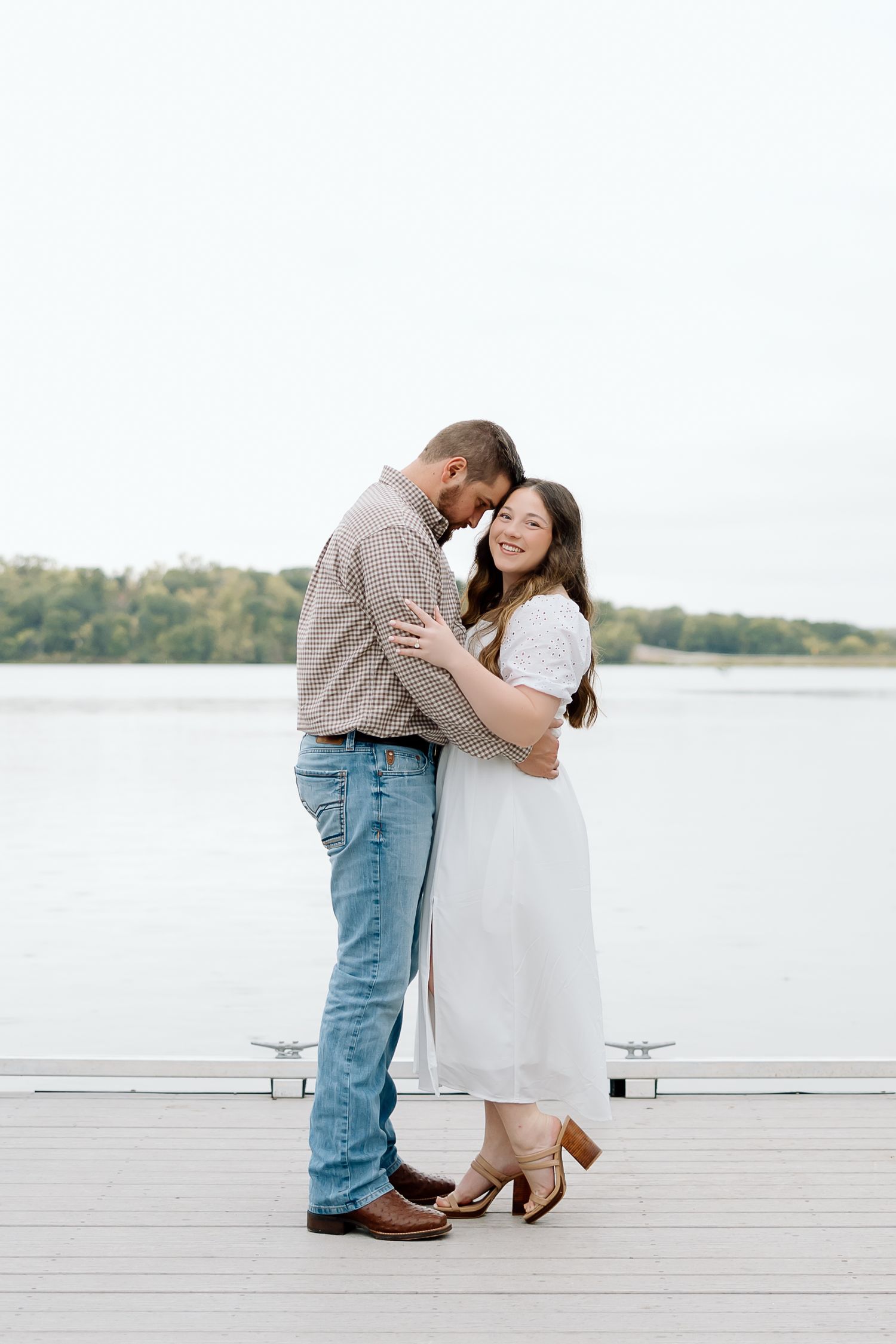 A couple laughs together while embracing on a wooden dock overlooking a lake.