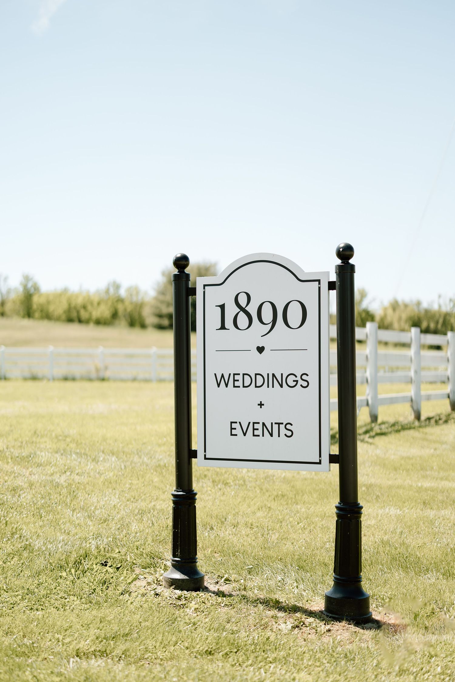 Elegant black and white wedding venue sign displaying '1890 Weddings & Events' on a sunny day with white fence.