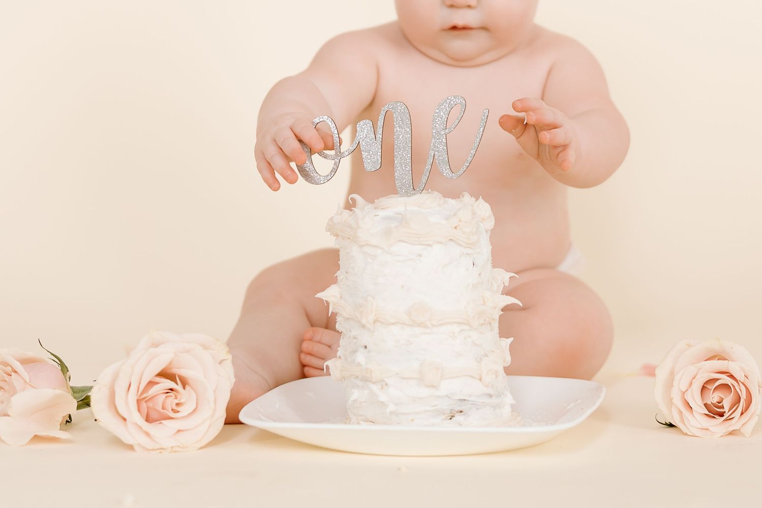 A baby reaches for a white birthday cake with 'one' topper surrounded by pink roses.