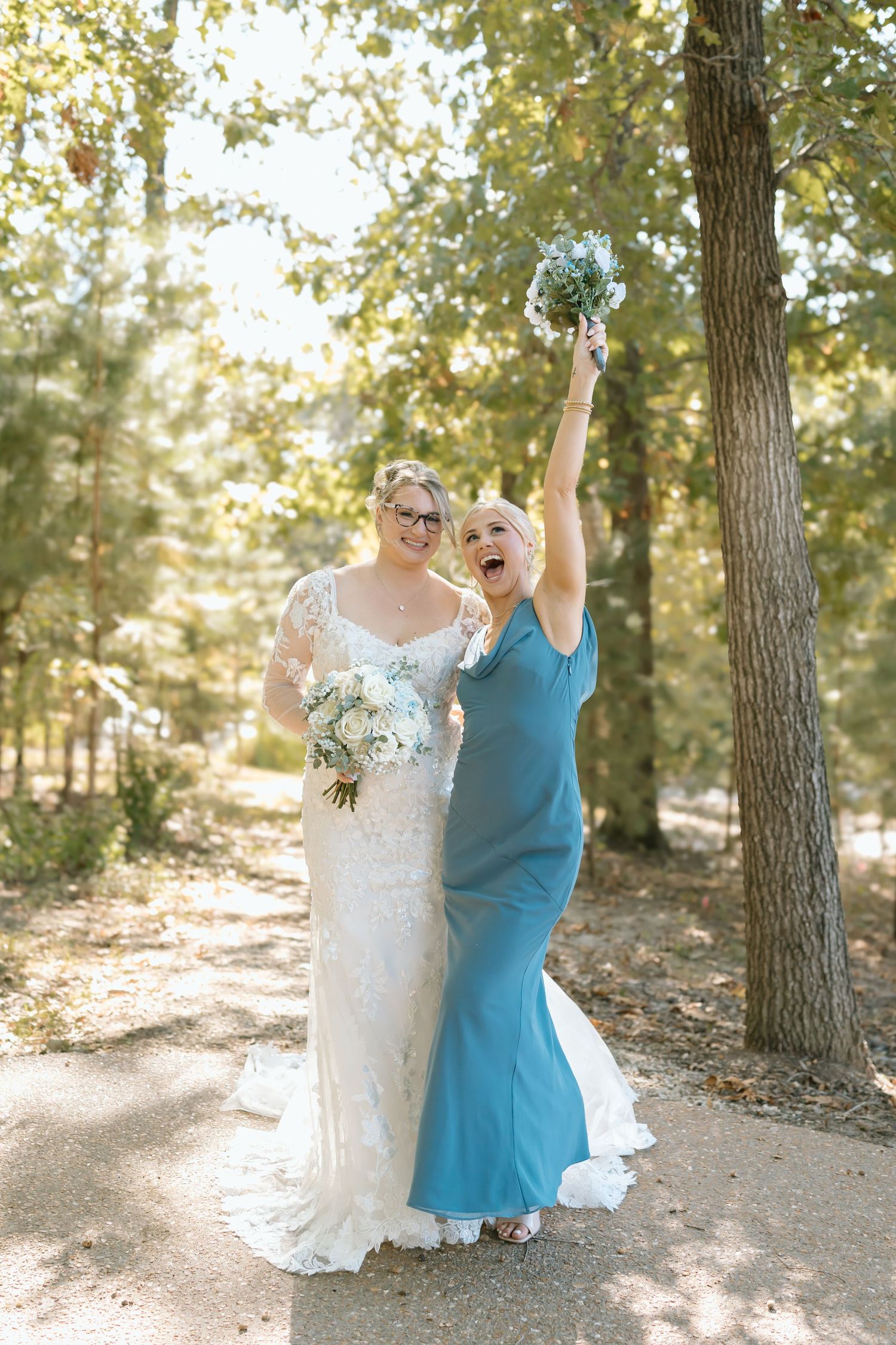 Two people in a wooded setting, one wearing a white dress and one in a turquoise dress, celebrating joyfully together.