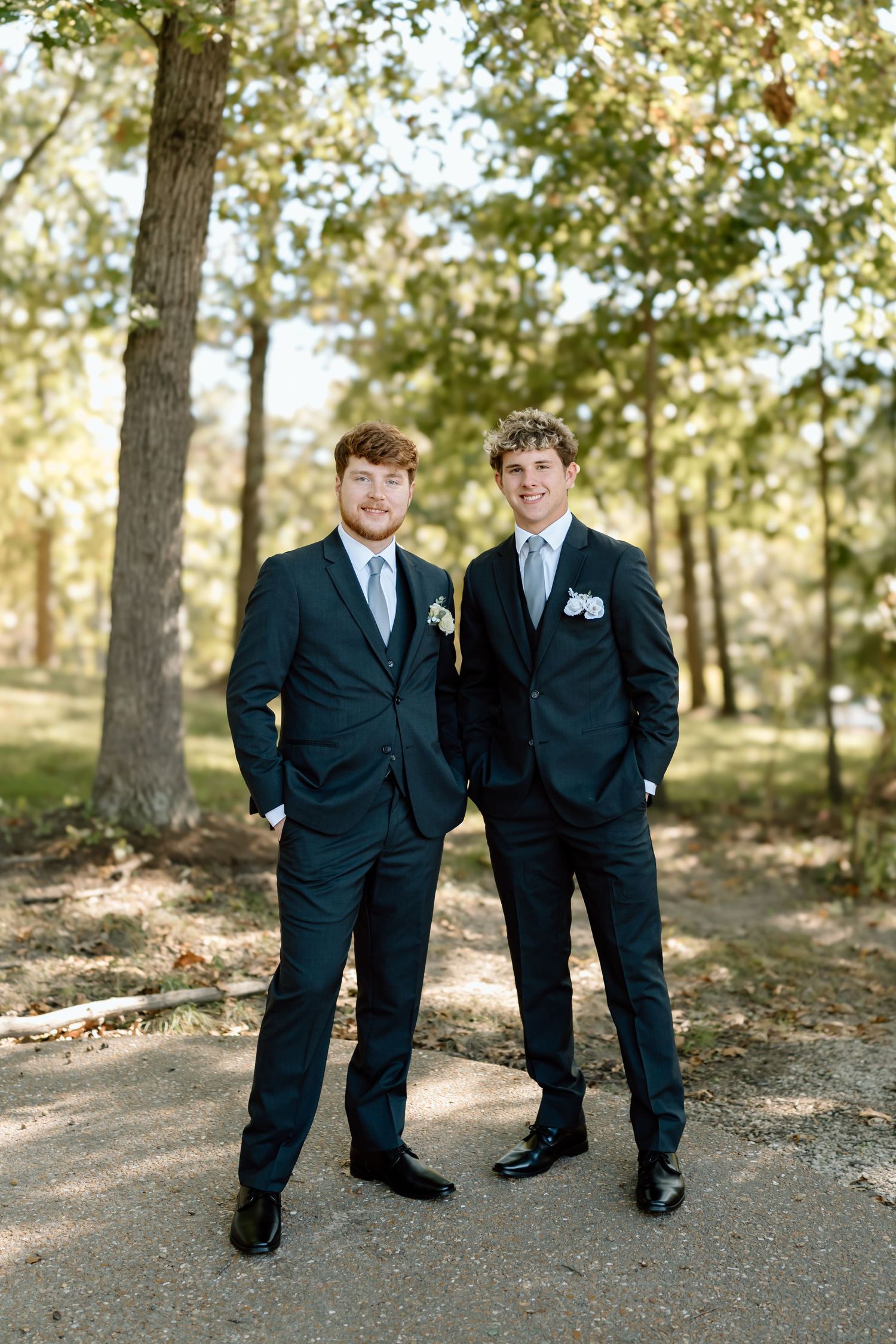 Two people in dark suits stand together on a wooded path during what appears to be a formal event or wedding.