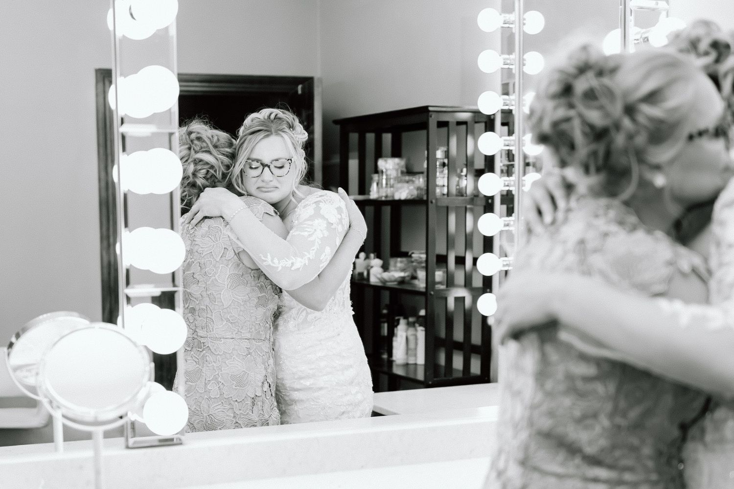 A black and white photo shows a person in lace getting ready at a vanity mirror with bright makeup lights.