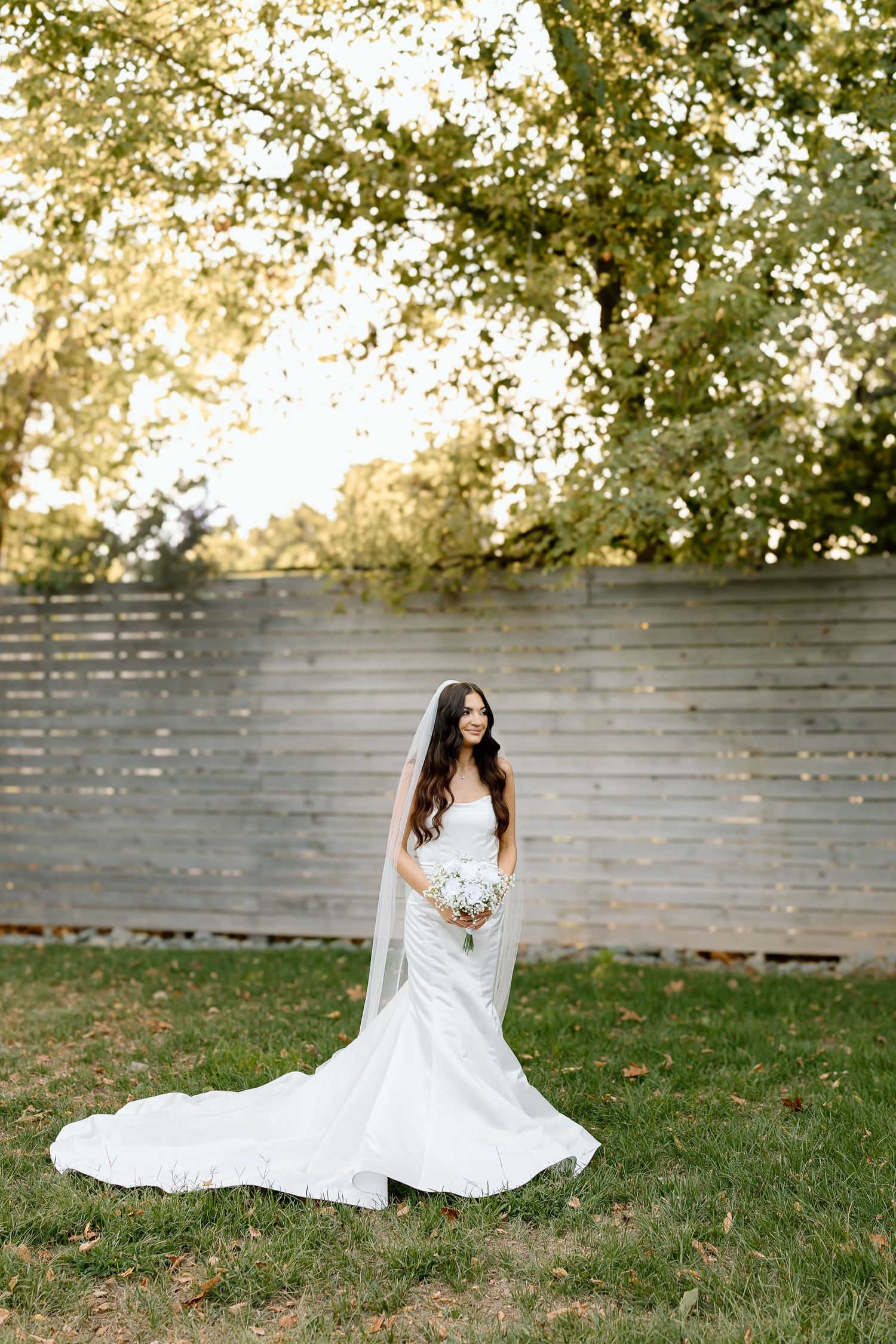 A flowing white wedding dress spins gracefully on a grassy lawn with a wooden fence in the background.