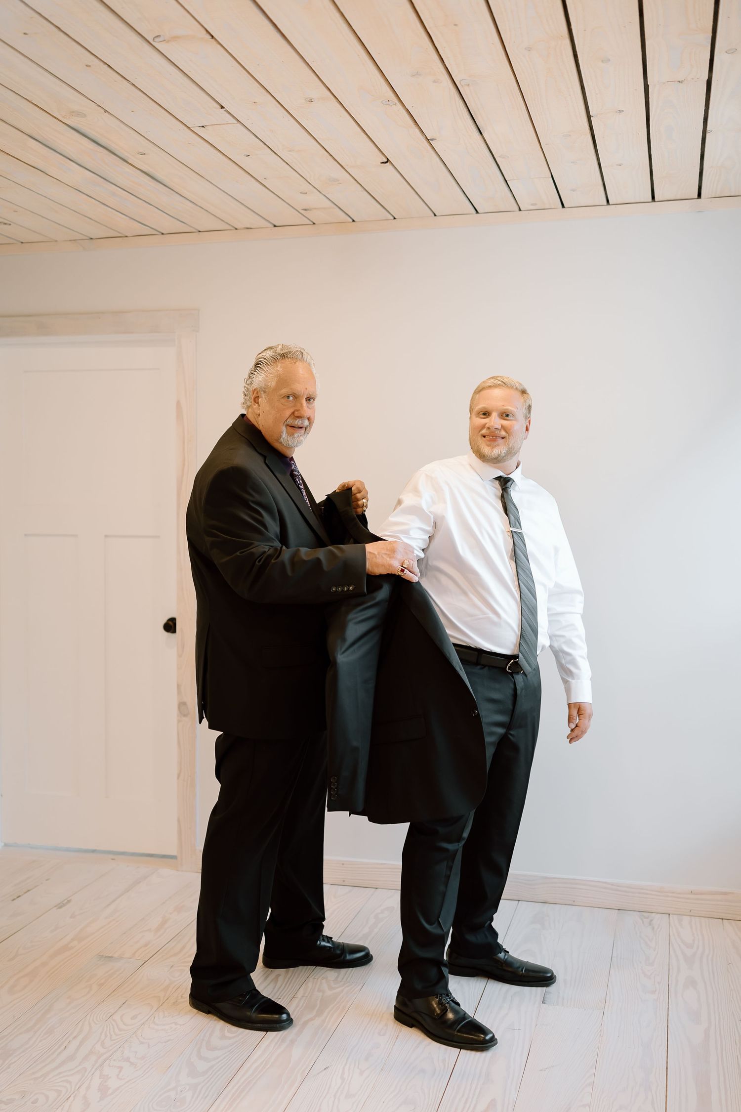 Two people in contrasting black and white formal attire stand together in a minimalist white room with wood ceiling.