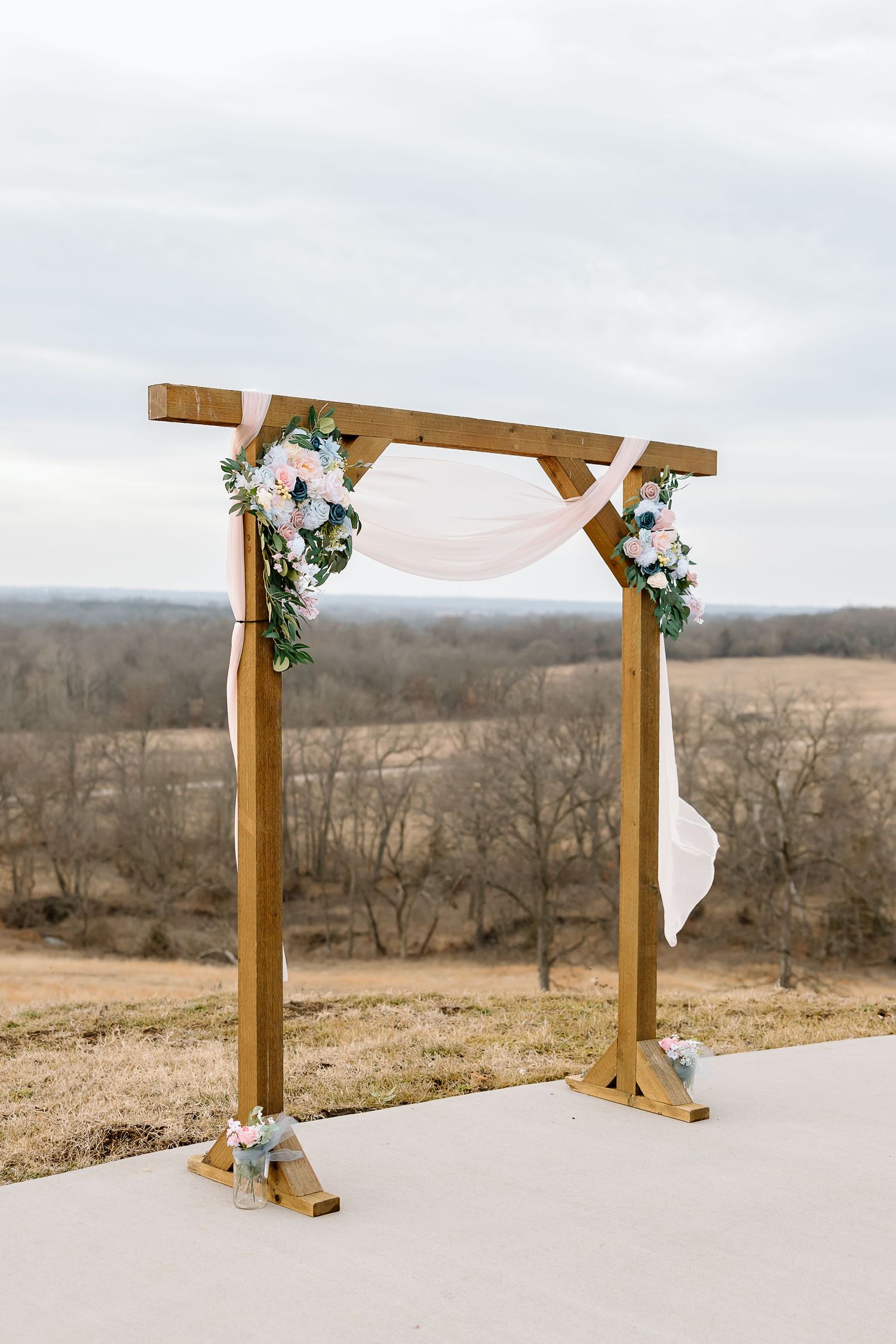 A rustic wooden wedding arch decorated with white fabric and floral arrangements stands against a scenic countryside backdrop.