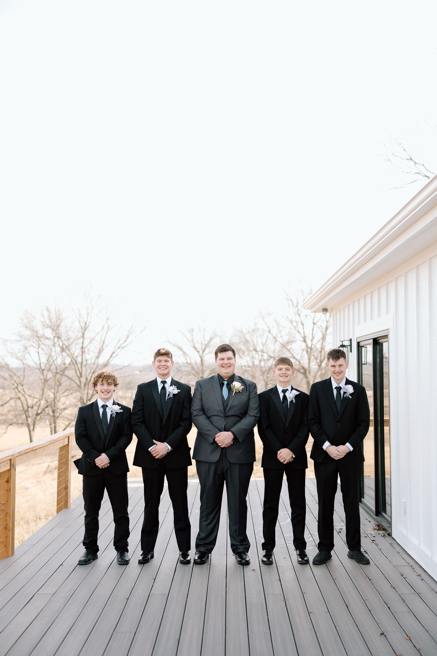 A group of five groomsmen in black suits stand in a row on a wooden deck outside a white building.