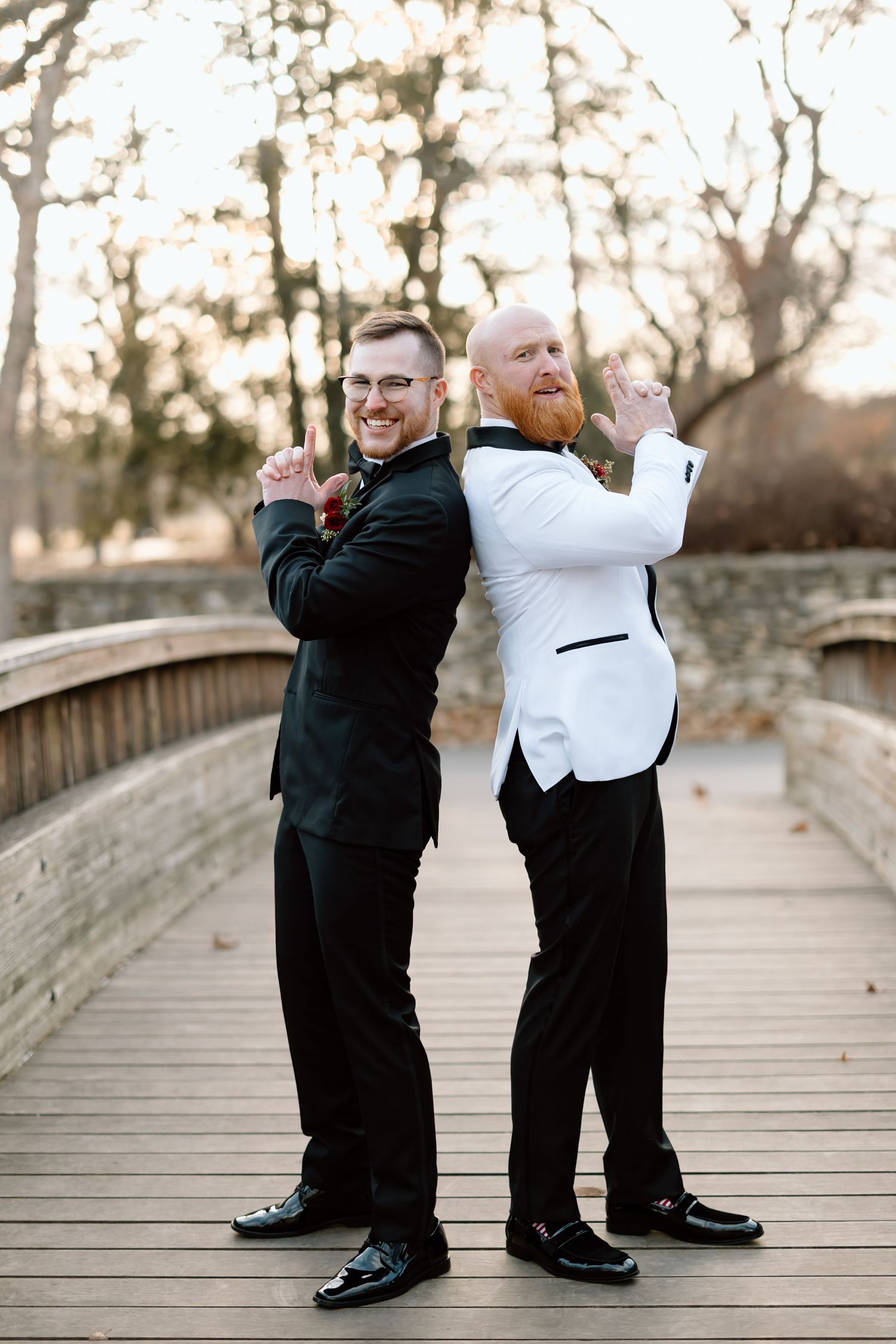 Two men in formal tuxedos pose back-to-back on a wooden bridge during sunset.