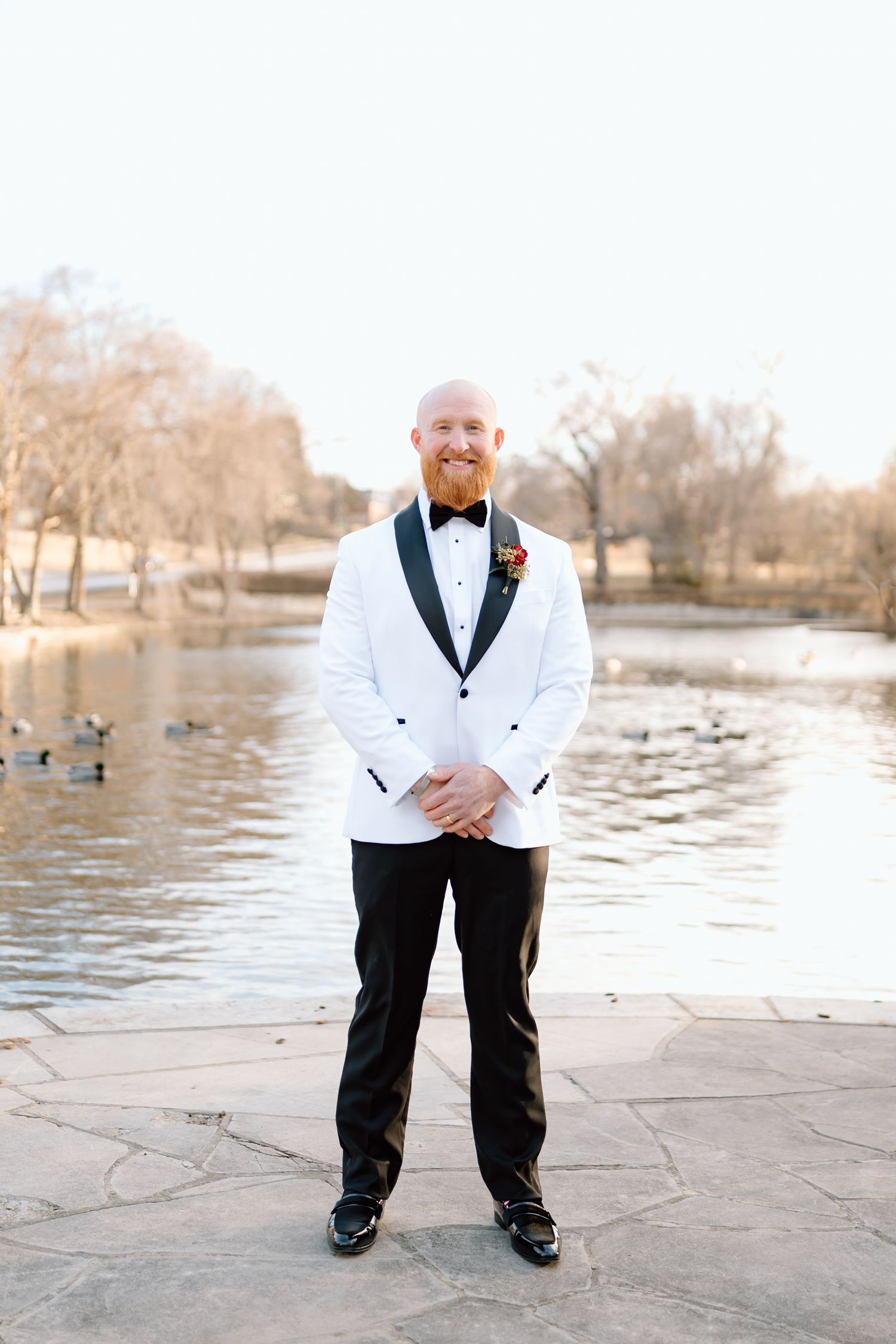 A groom in a white tuxedo jacket and bow tie poses by a lake with ducks swimming in the background.