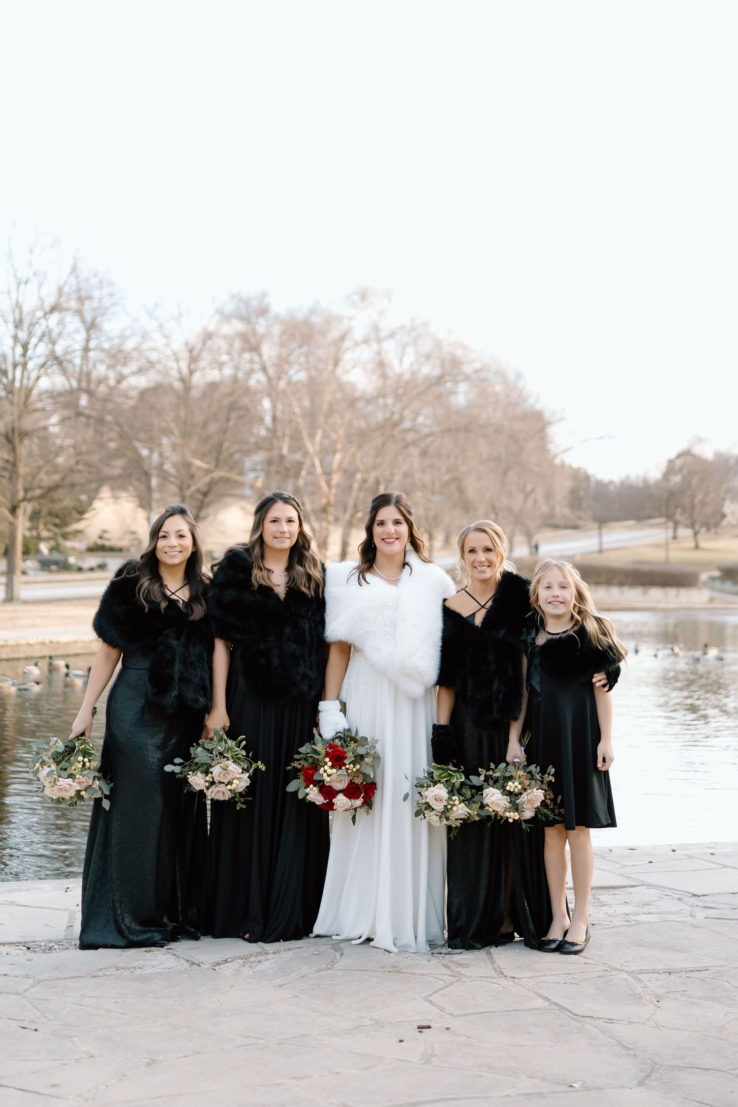Wedding party in black dresses and white gown pose together by a lakeside on a winter day.