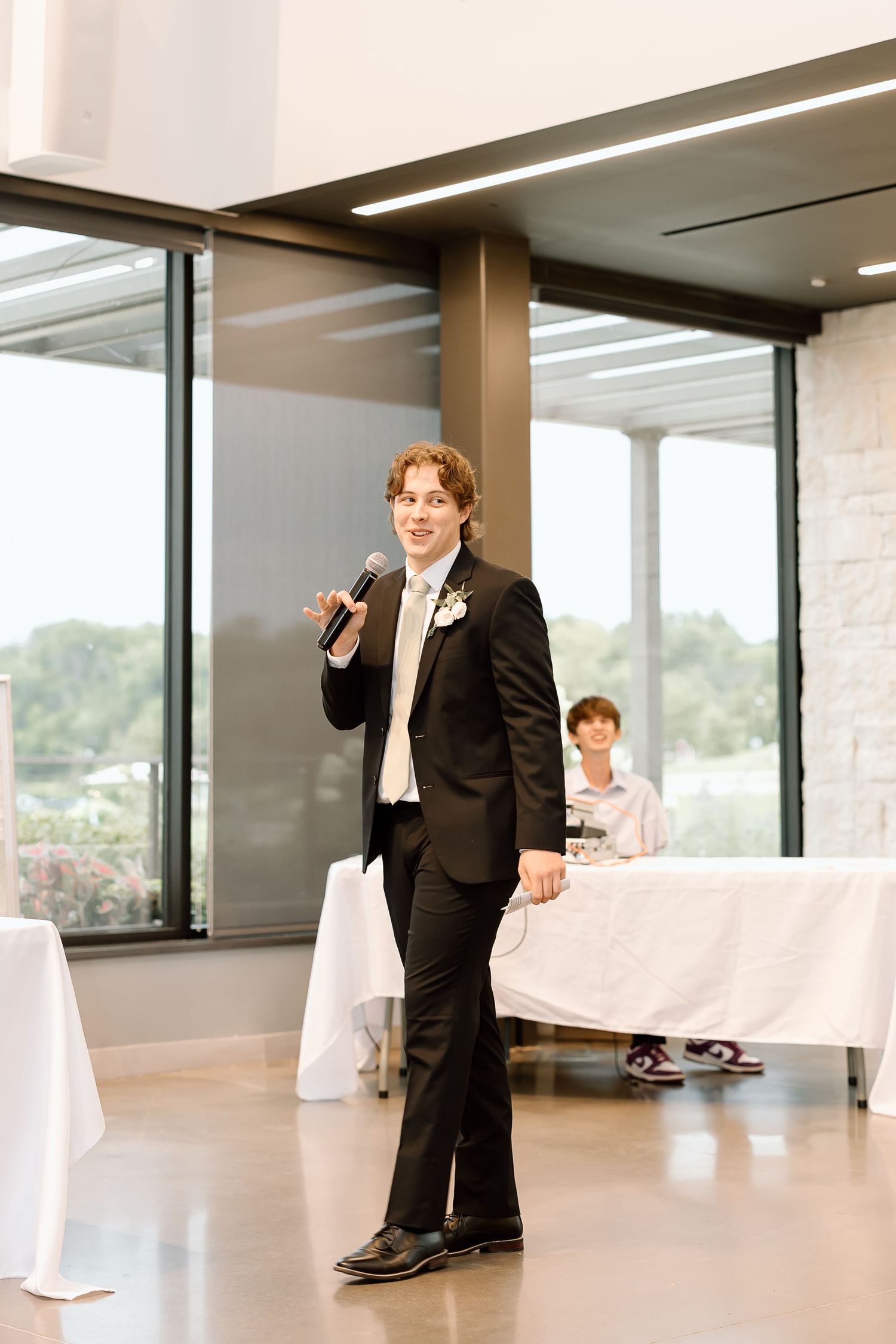 A person in a black tuxedo speaks into a microphone at an elegant event with white tablecloths and large windows.