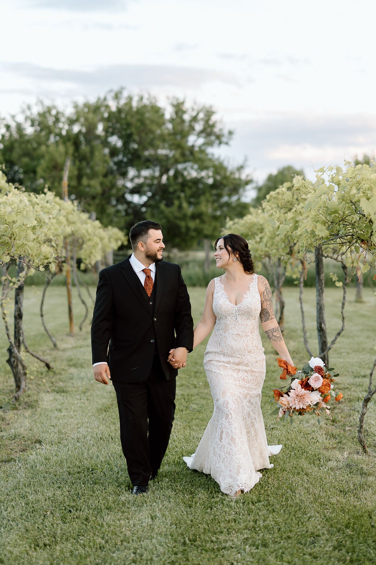 A bride and groom walk hand in hand through a vineyard at sunset, with the bride holding a colorful autumn bouquet.