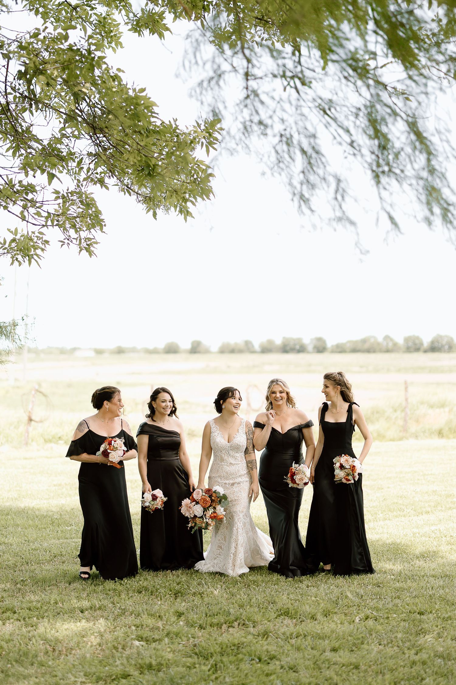 A wedding party stands together on a grassy field beneath overhanging tree branches on a sunny day.