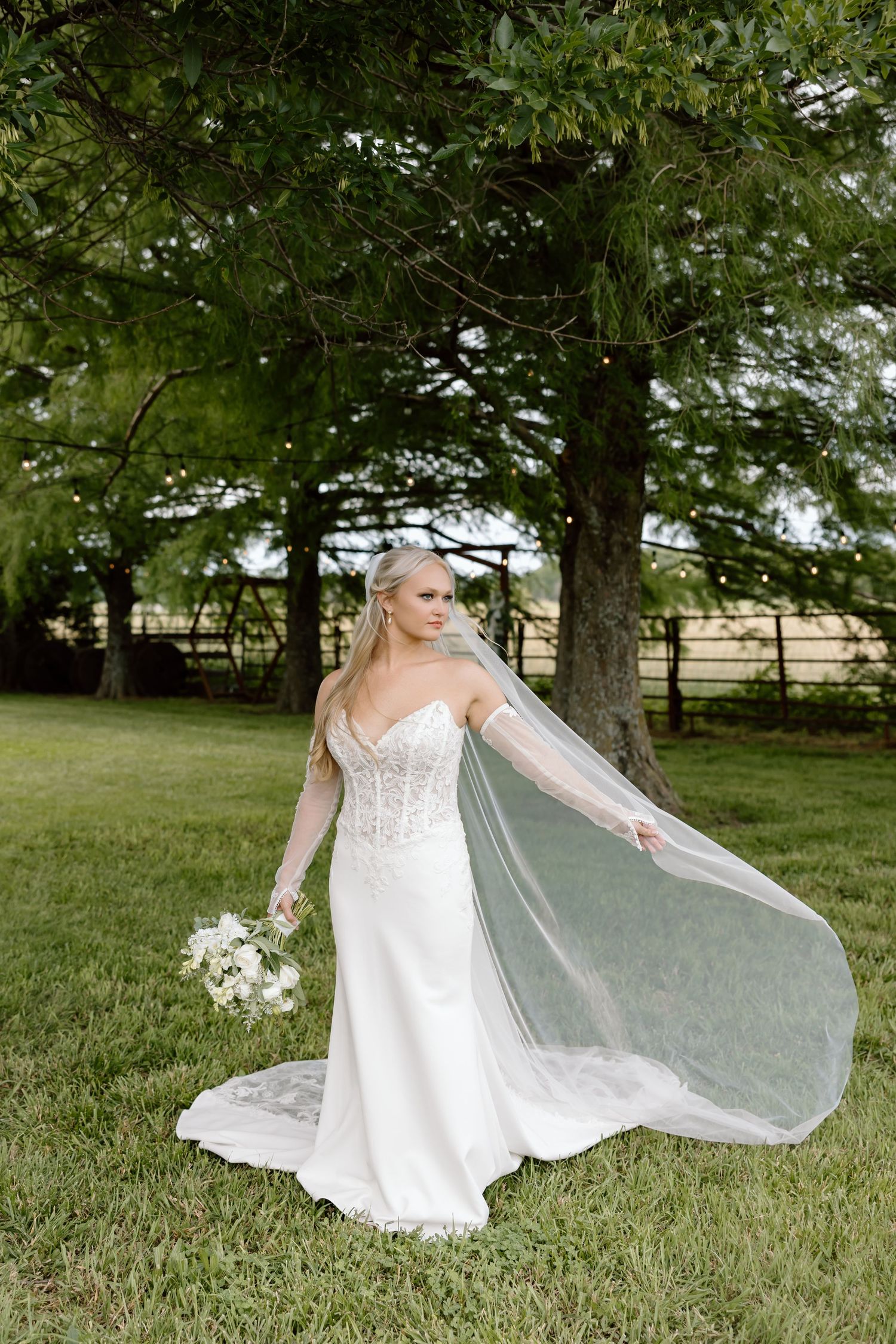 Elegant bride in lace wedding gown holding white bouquet with cathedral veil flowing outdoors near large tree.