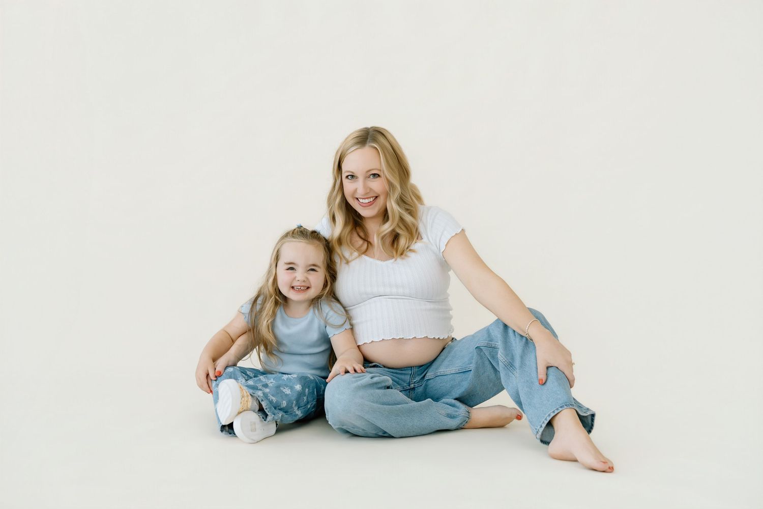 Pregnant woman and young daughter sitting together smiling on white background in matching casual outfits.