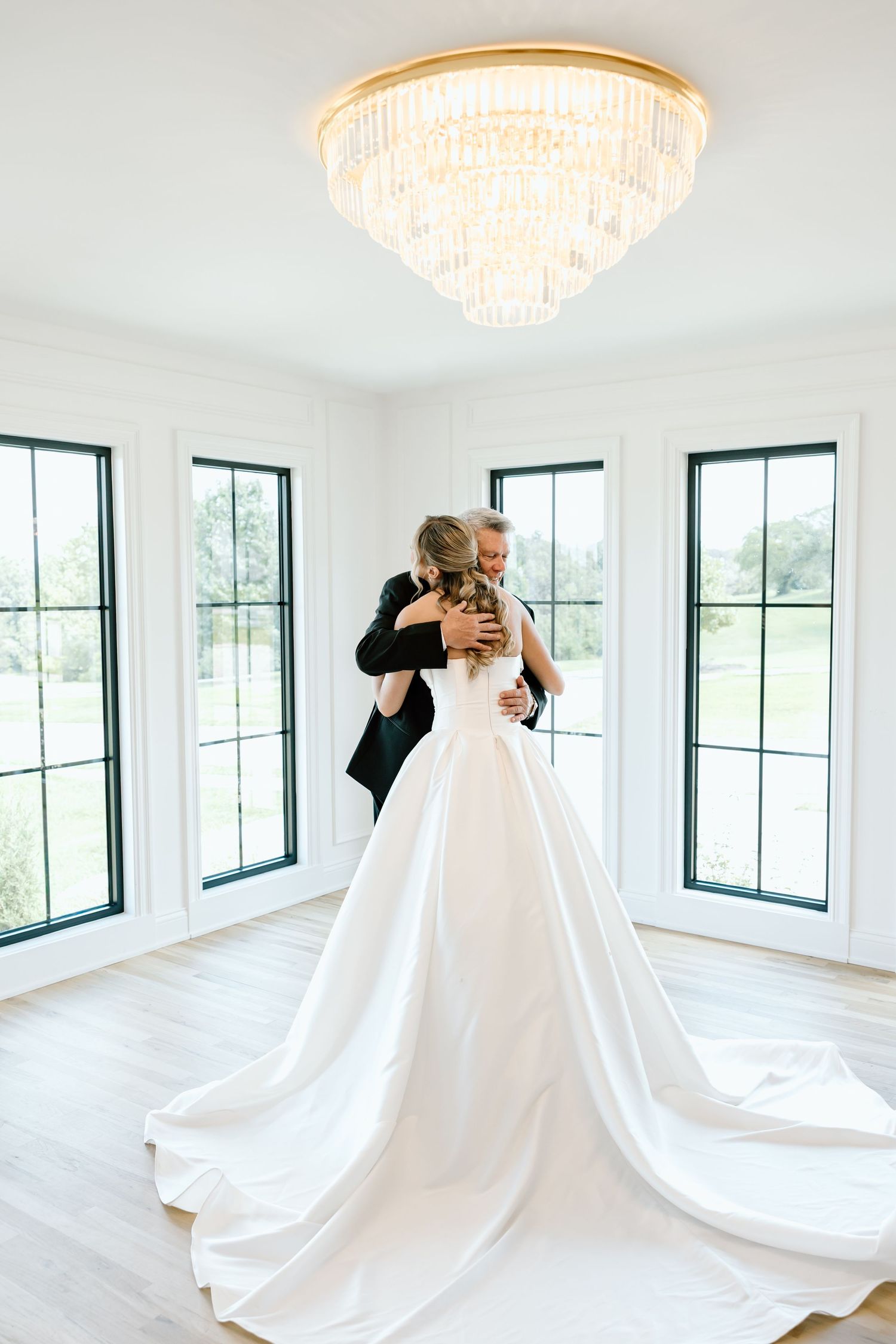 Bride and groom share a tender embrace in elegant white room with crystal chandelier and large windows on wedding day.