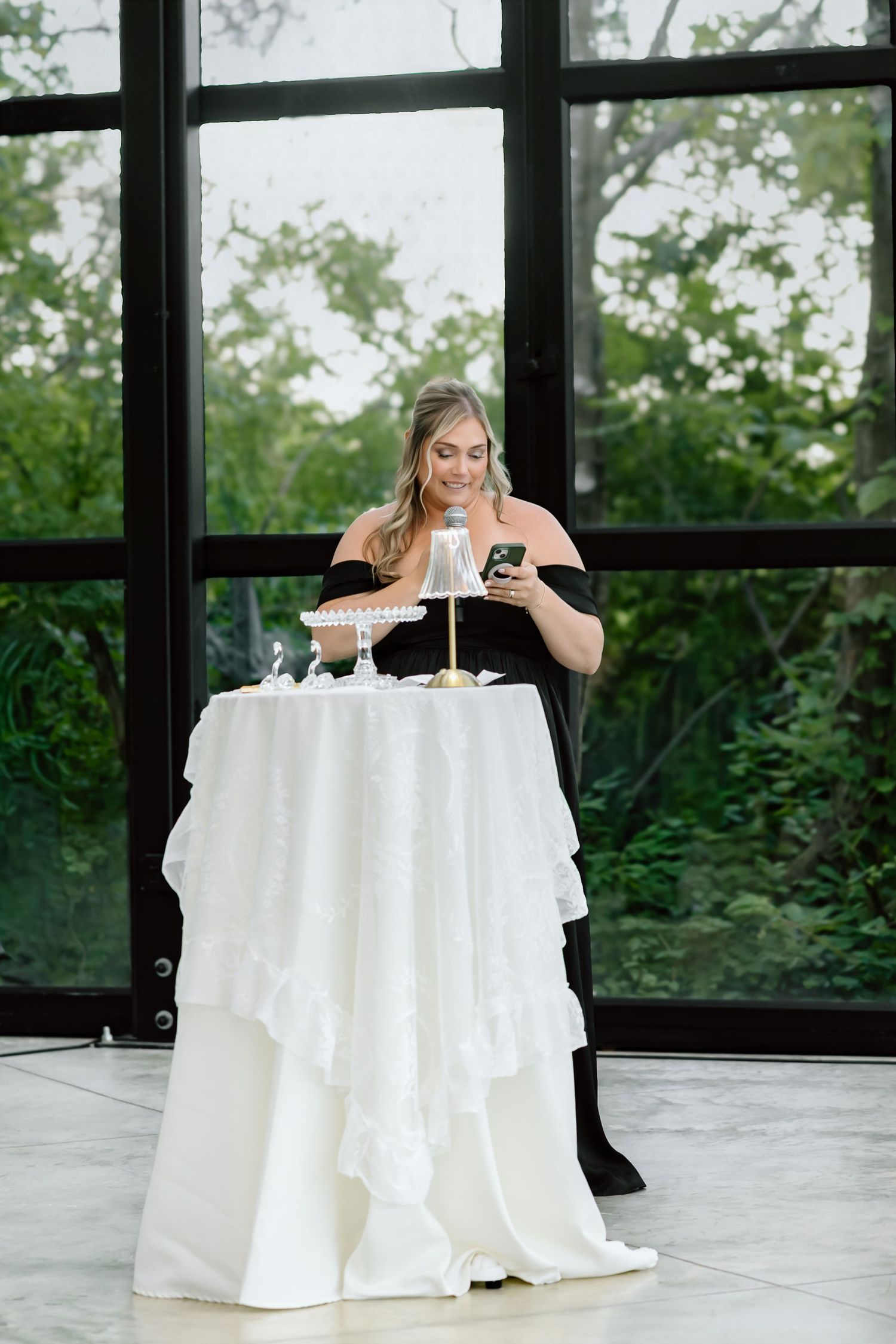 Woman in black dress smiling at phone beside elegant white ruffled cocktail table at indoor event.