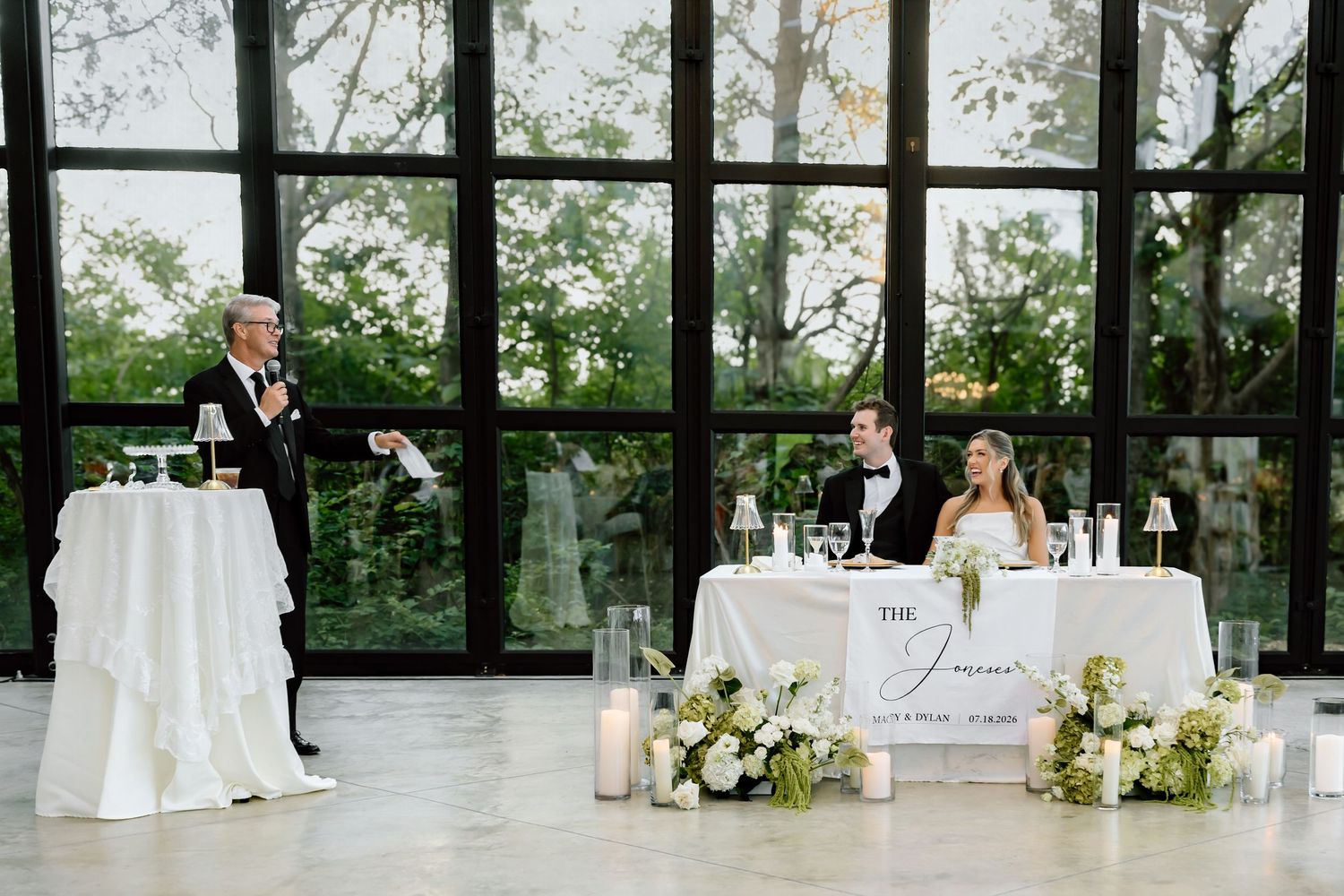Wedding reception toast with groom at podium and bride seated at sweetheart table, surrounded by candles and white floral arrangements.