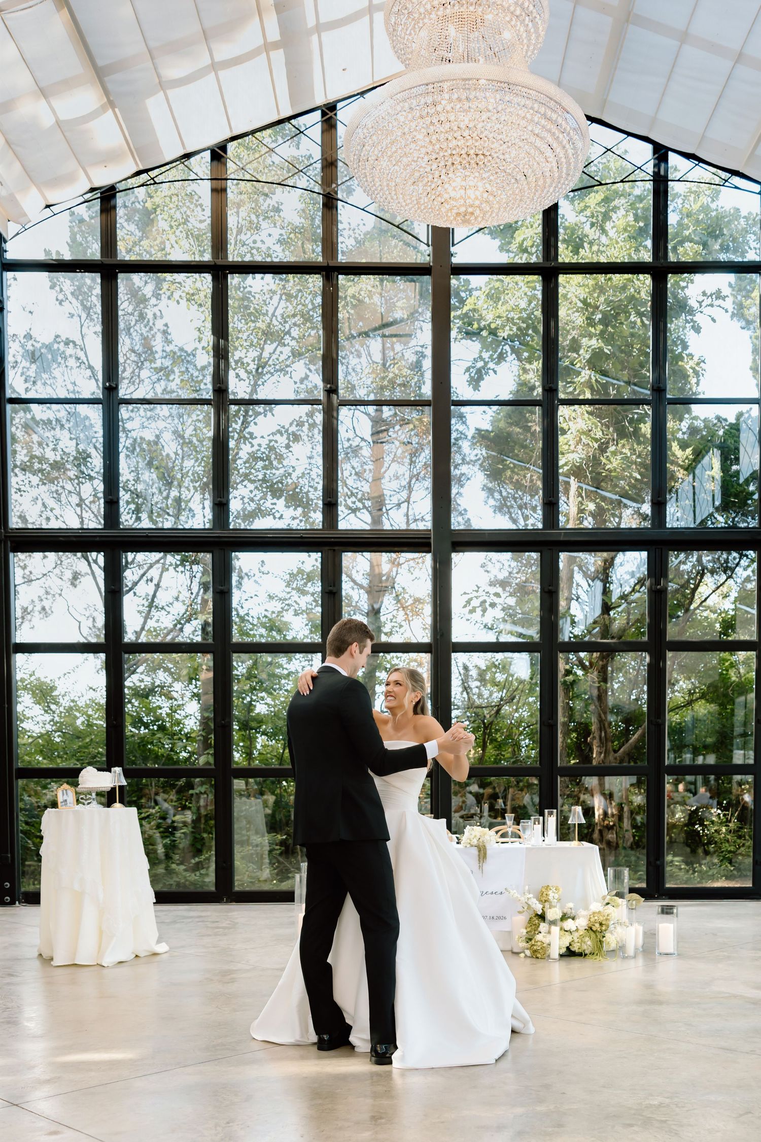 Bride and groom share first dance in elegant glass-walled venue with crystal chandelier and lush garden views.