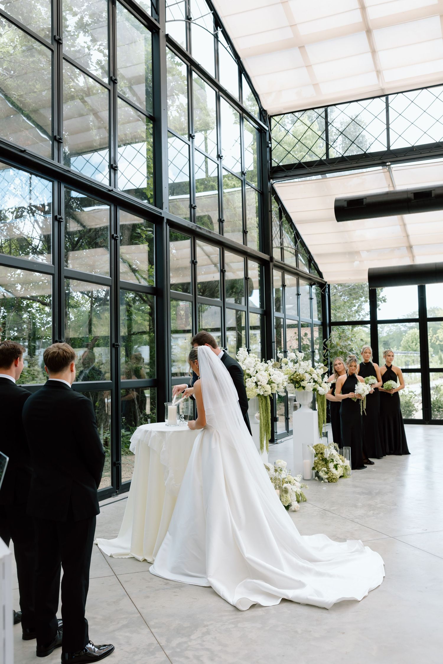 Bride and groom at altar in elegant glass greenhouse wedding venue with black-clad bridesmaids and white floral arrangements.