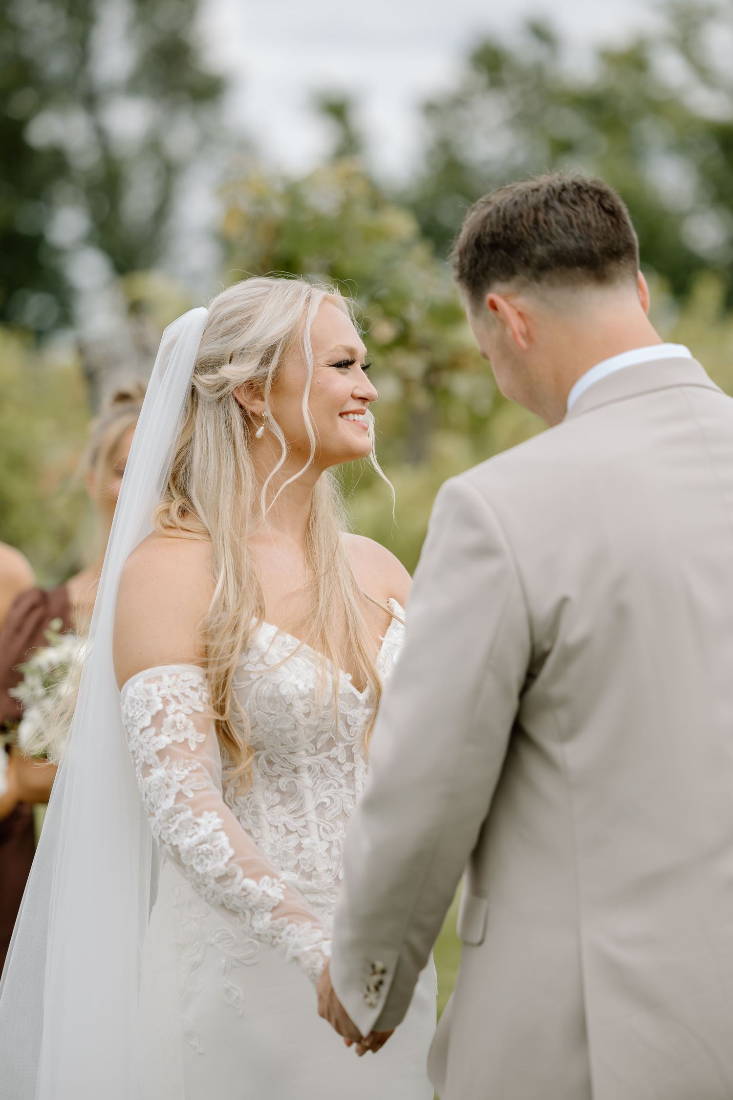 Smiling bride in lace off-shoulder gown and veil holds hands with groom in tan suit during outdoor wedding ceremony.