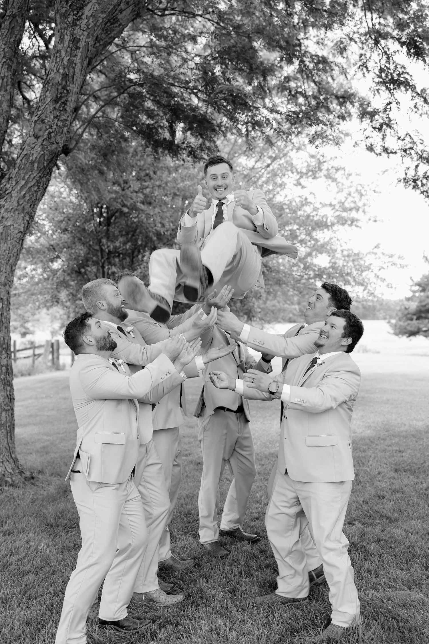 Groomsmen tossing groom in the air outdoors in matching suits, black and white wedding photo.
