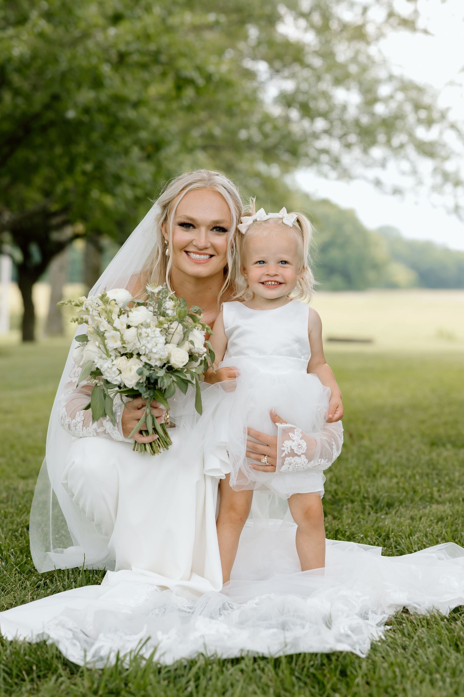 Bride in white gown holding bouquet poses with smiling toddler flower girl on green lawn.