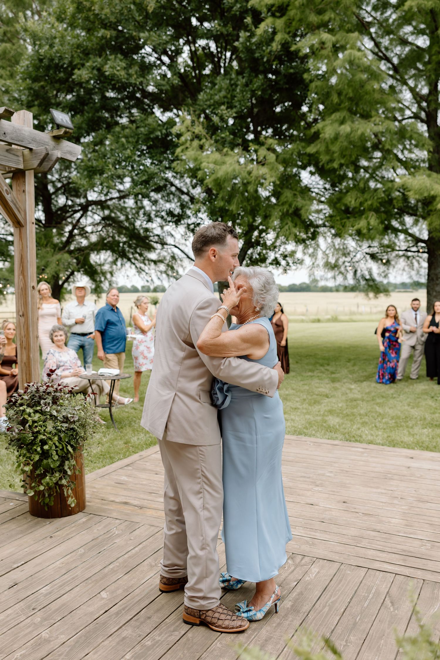 Groom shares a tender dance with elderly woman in light blue dress at outdoor rustic wedding reception on wooden deck.