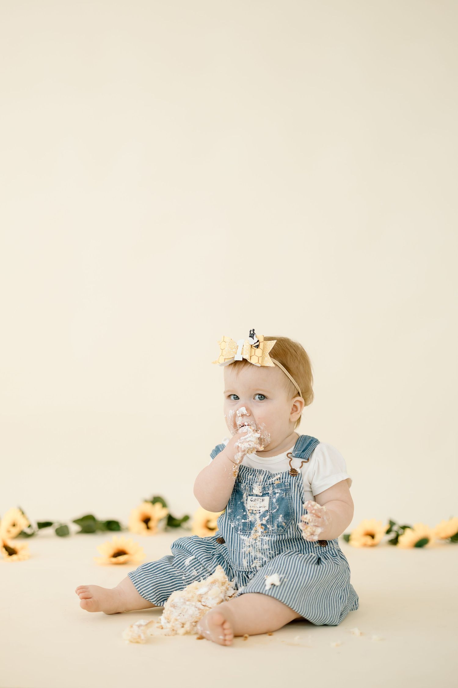 A baby in denim overalls sits on a cream backdrop with scattered flowers during a cake smash photo session.