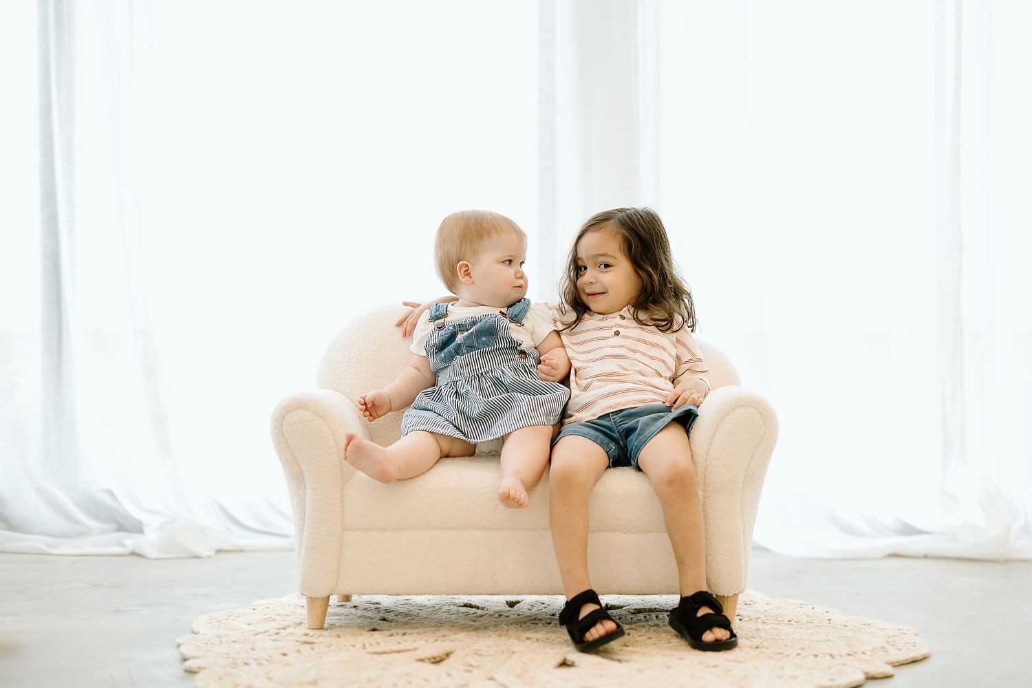 Two young children sit together on a beige couch in a bright white room with sheer curtains.