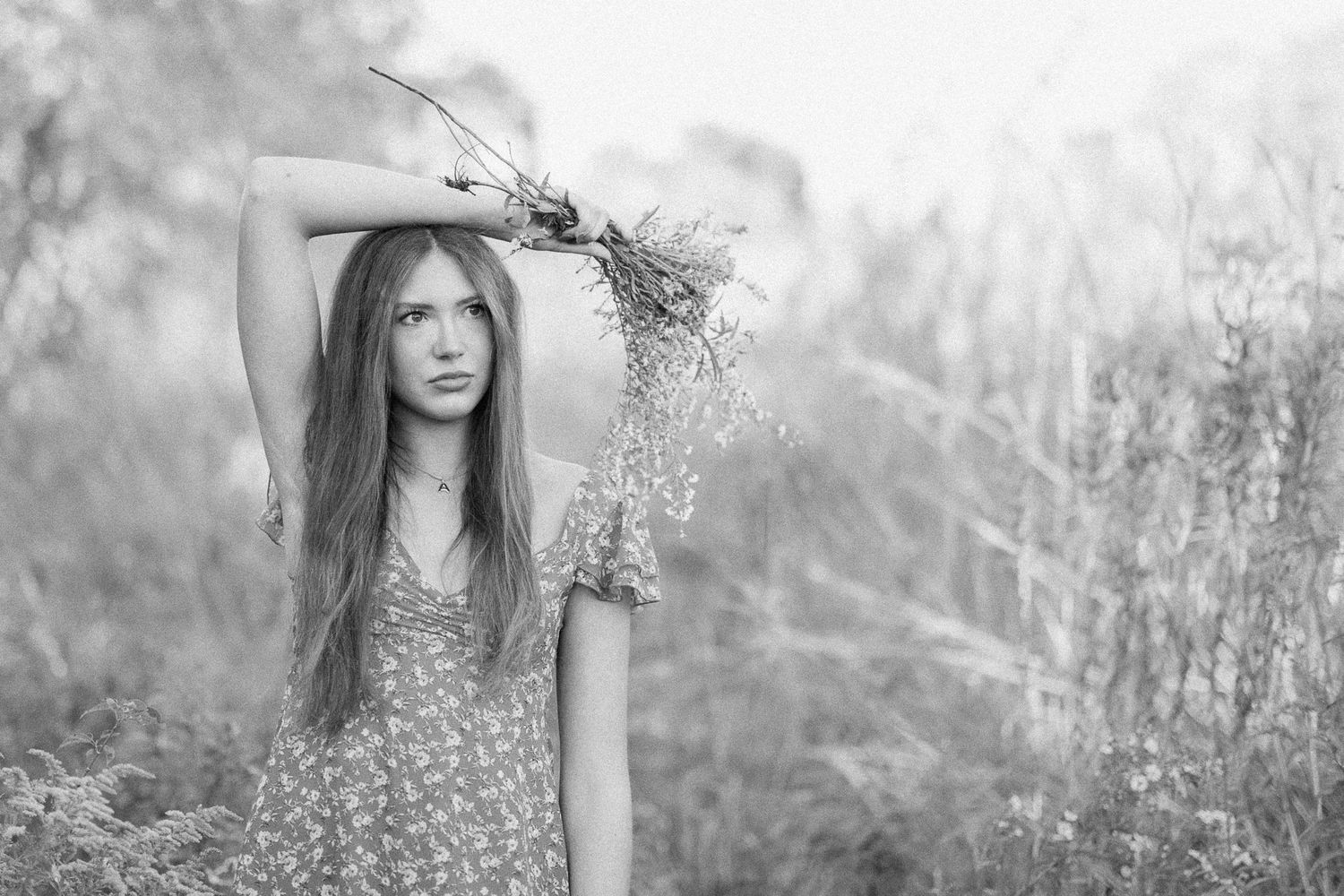 Black and white portrait of a person with long hair standing in a field of tall grass and wildflowers.