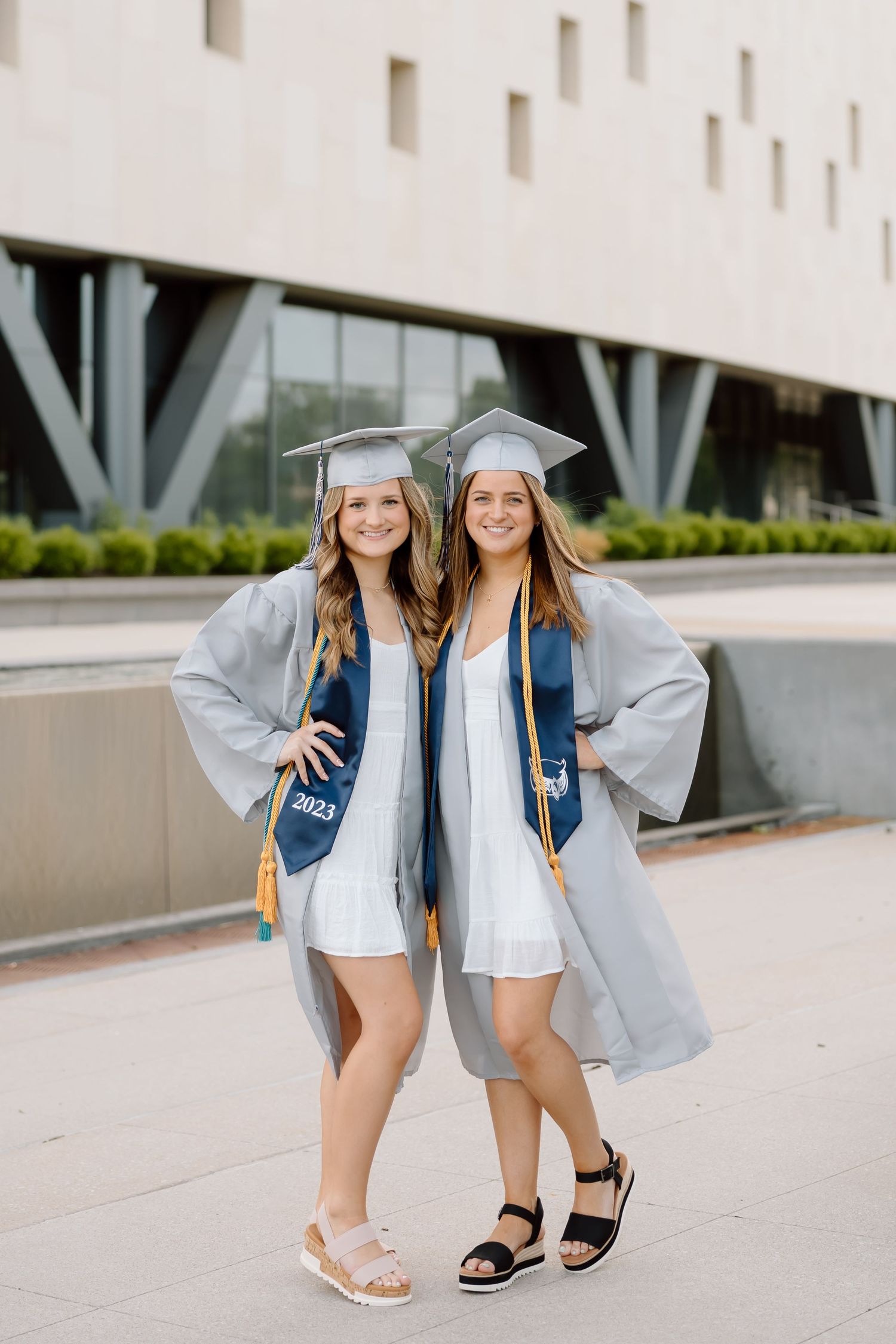 Two graduates in silver gowns and blue stoles stand together on campus showing their matching outfits and sandals.