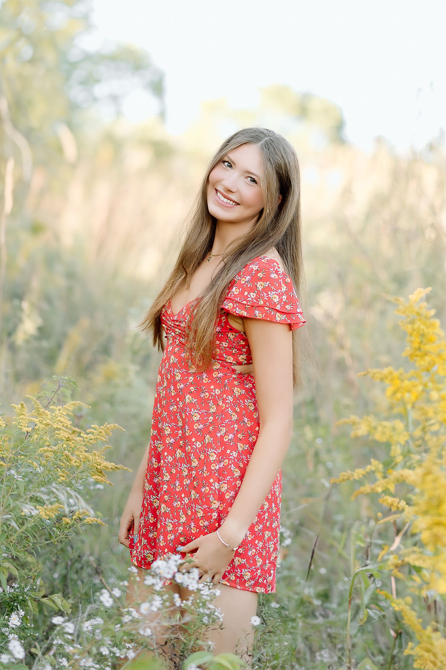 A person in a red floral dress poses in a meadow with yellow wildflowers during golden hour.