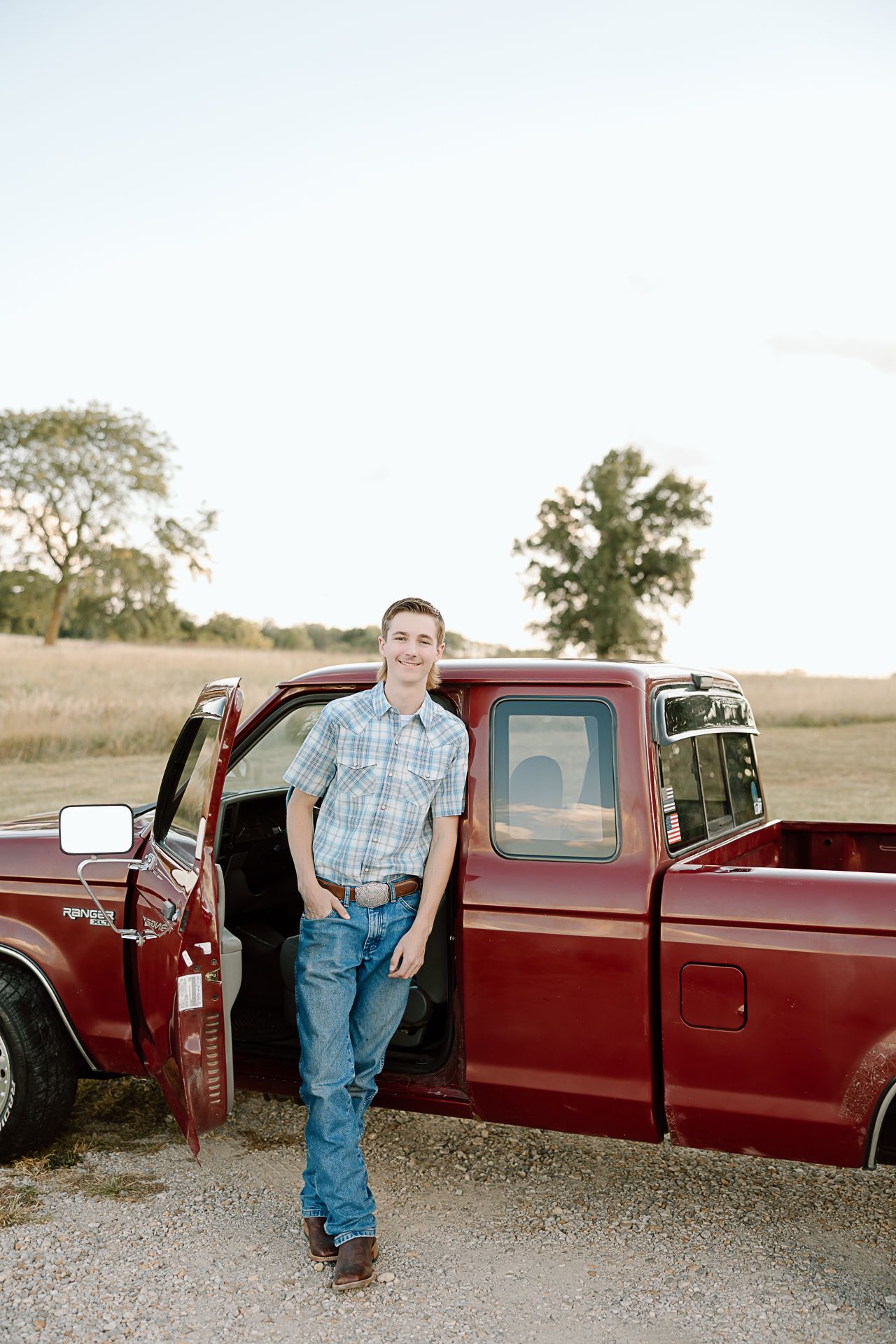 Someone poses casually while leaning against the door of a classic red pickup truck in a countryside setting.