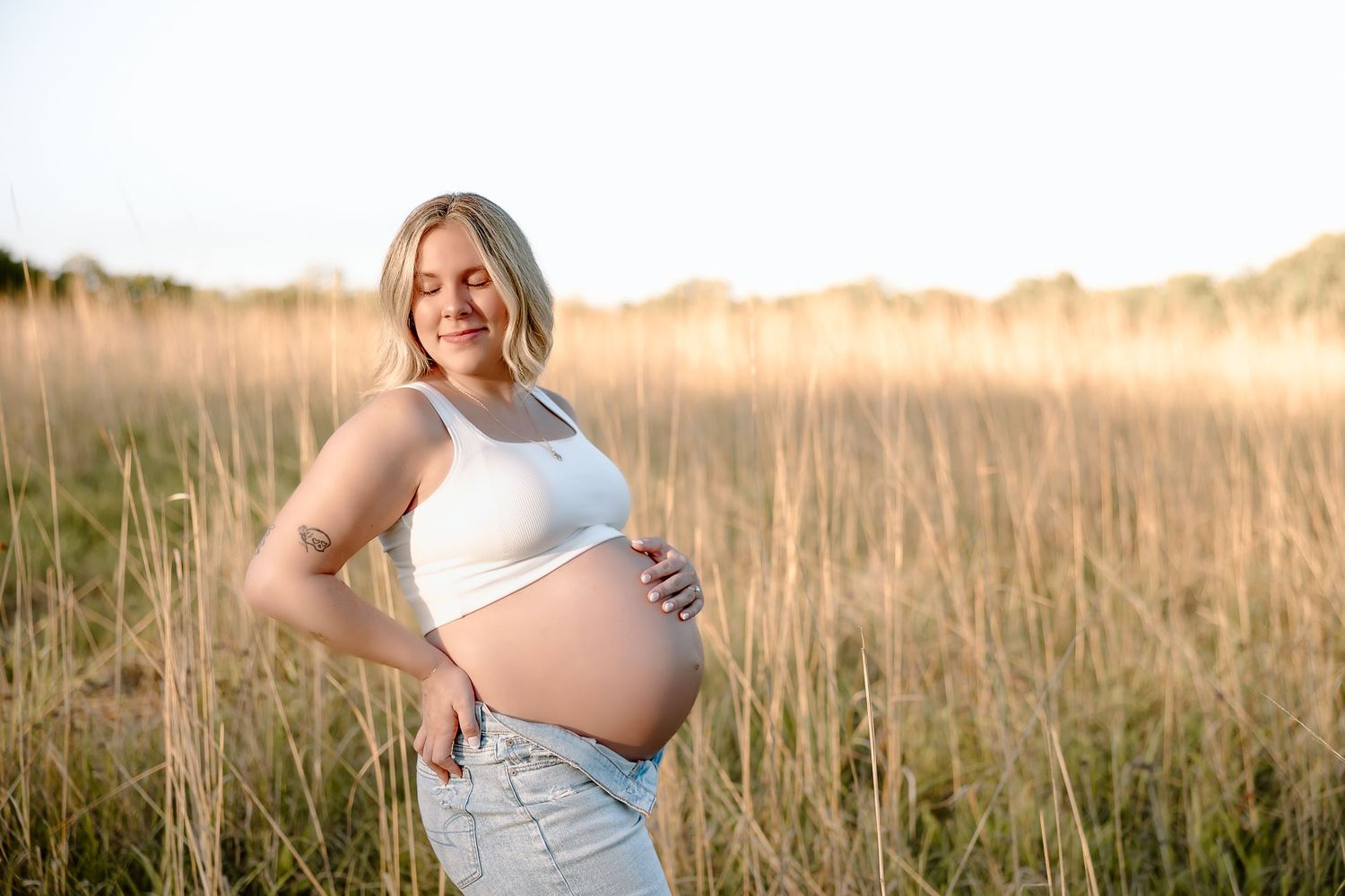 A person in a white top poses in a golden field during sunset, creating a serene maternity photoshoot scene.
