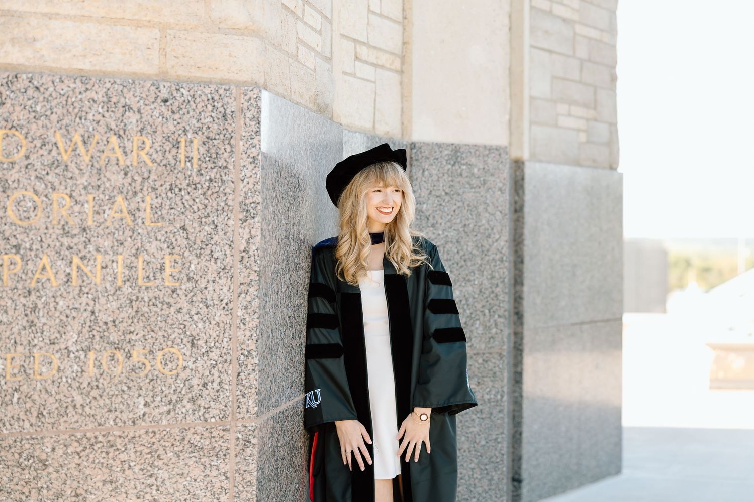Graduate in cap and gown poses against stone building wall during graduation photo session.