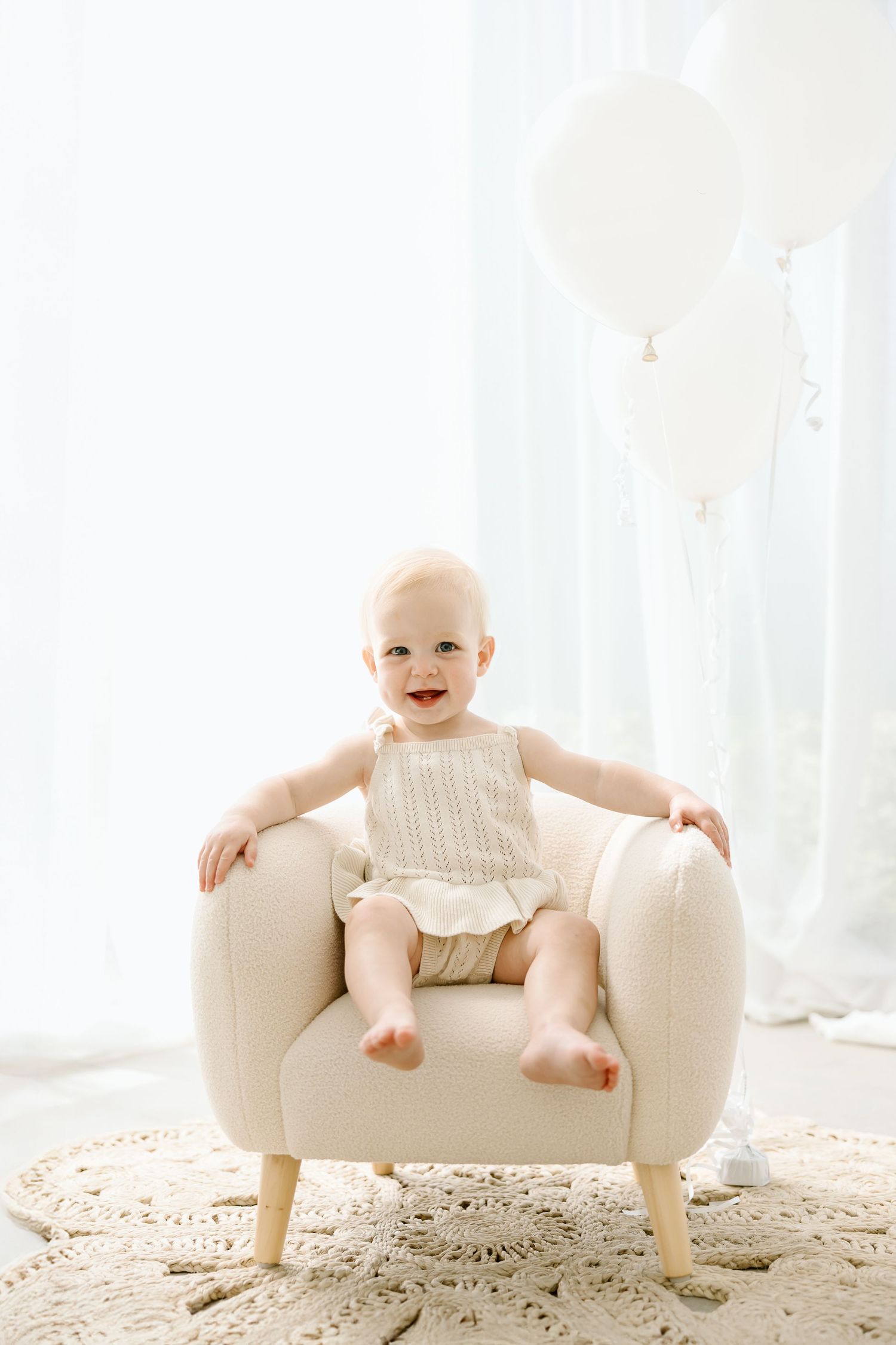 Happy baby wearing cream-colored outfit sits in cozy white armchair against bright window with sheer curtains.