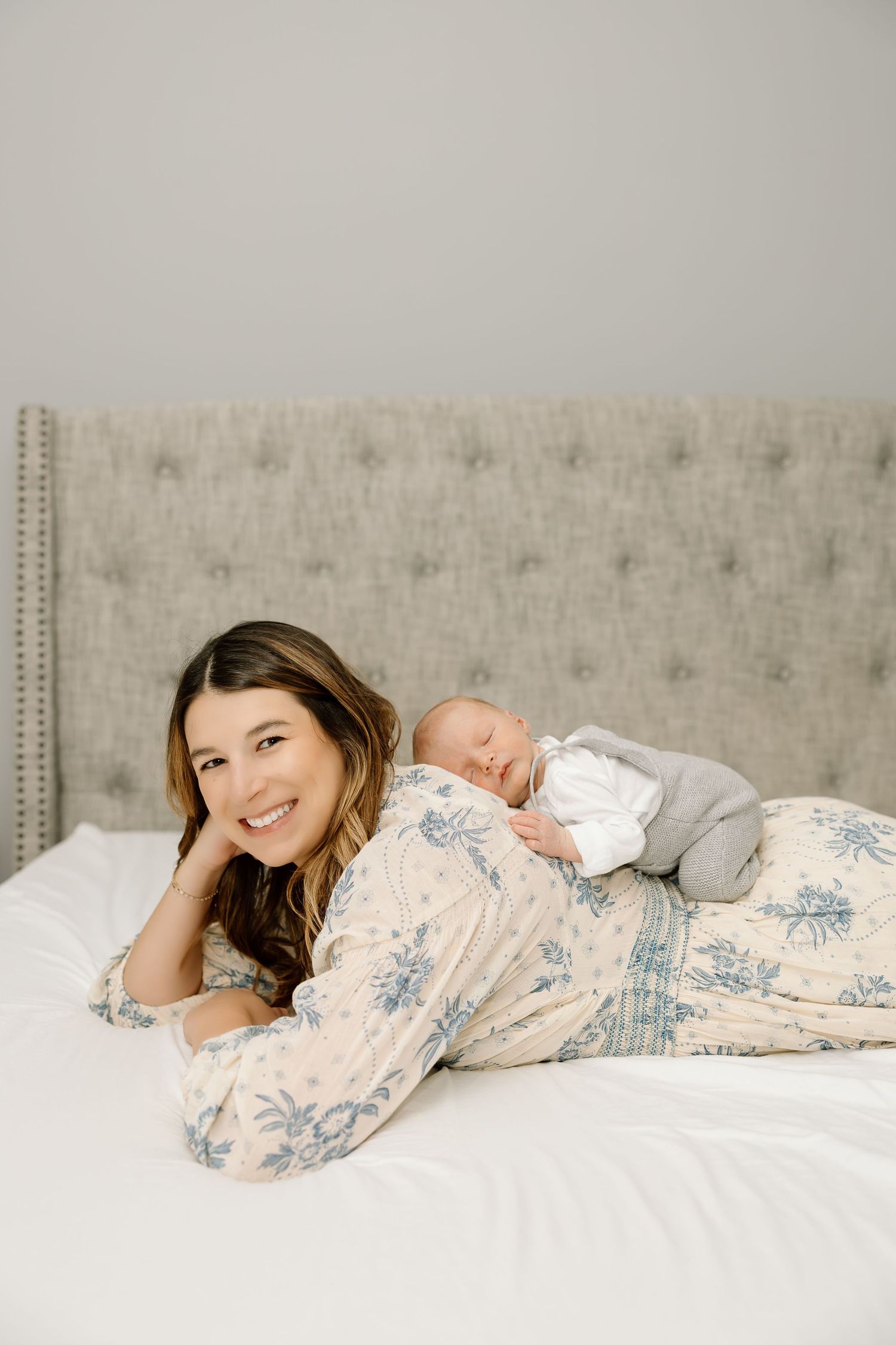 A person in a floral print dress lies comfortably on a tufted gray headboard bed with white bedding.