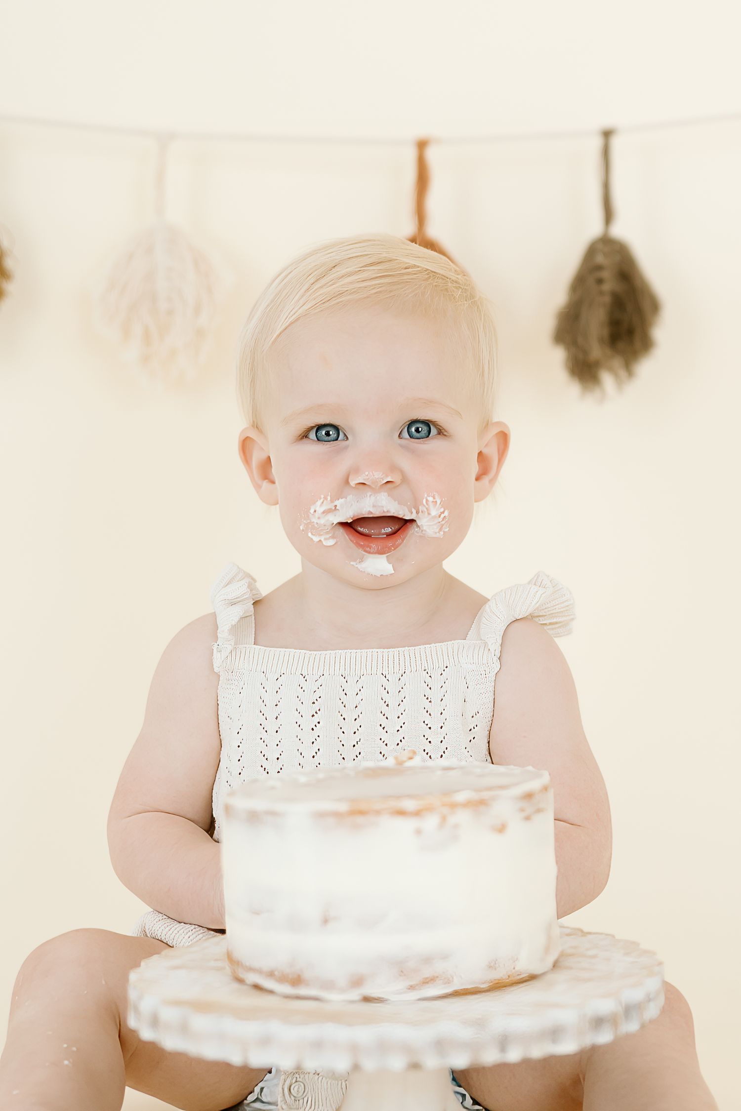 Baby in white lace dress smiles while sitting with birthday cake in front of rustic backdrop.