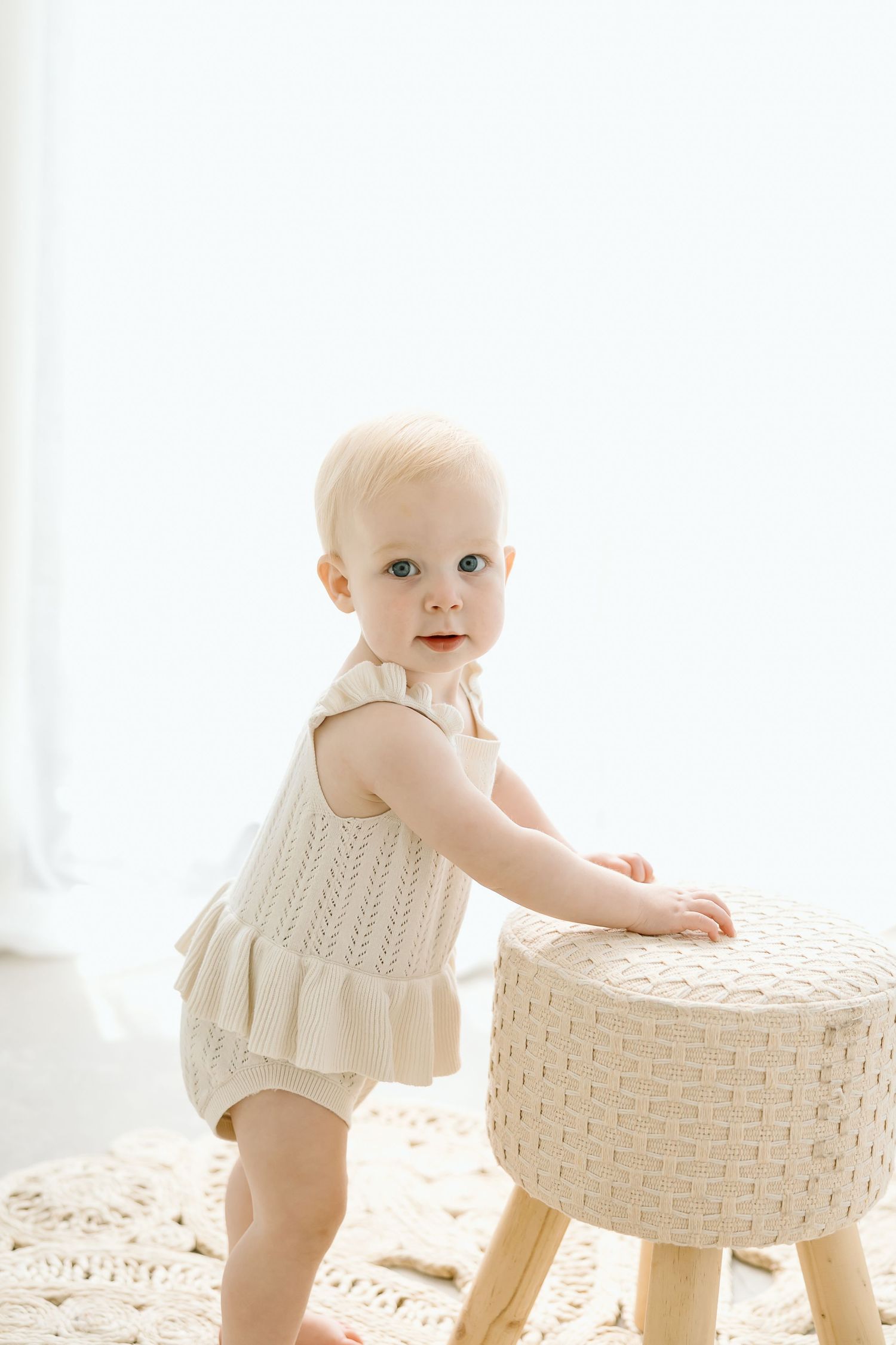 Baby in white ruffled outfit crawls around woven basket in bright sunlit room.