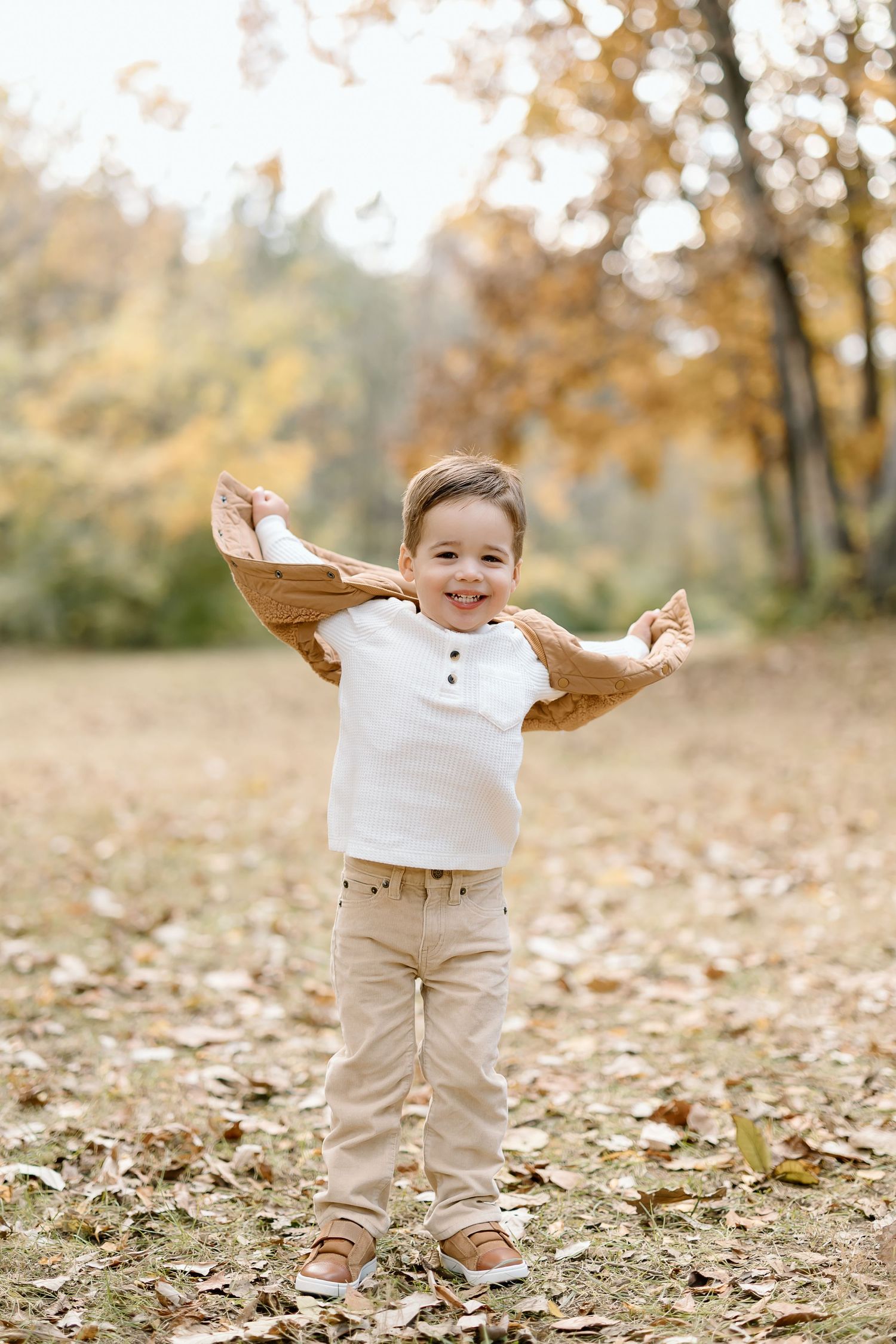 A child in beige pants and white sweater plays joyfully in fallen autumn leaves on a sunny day.