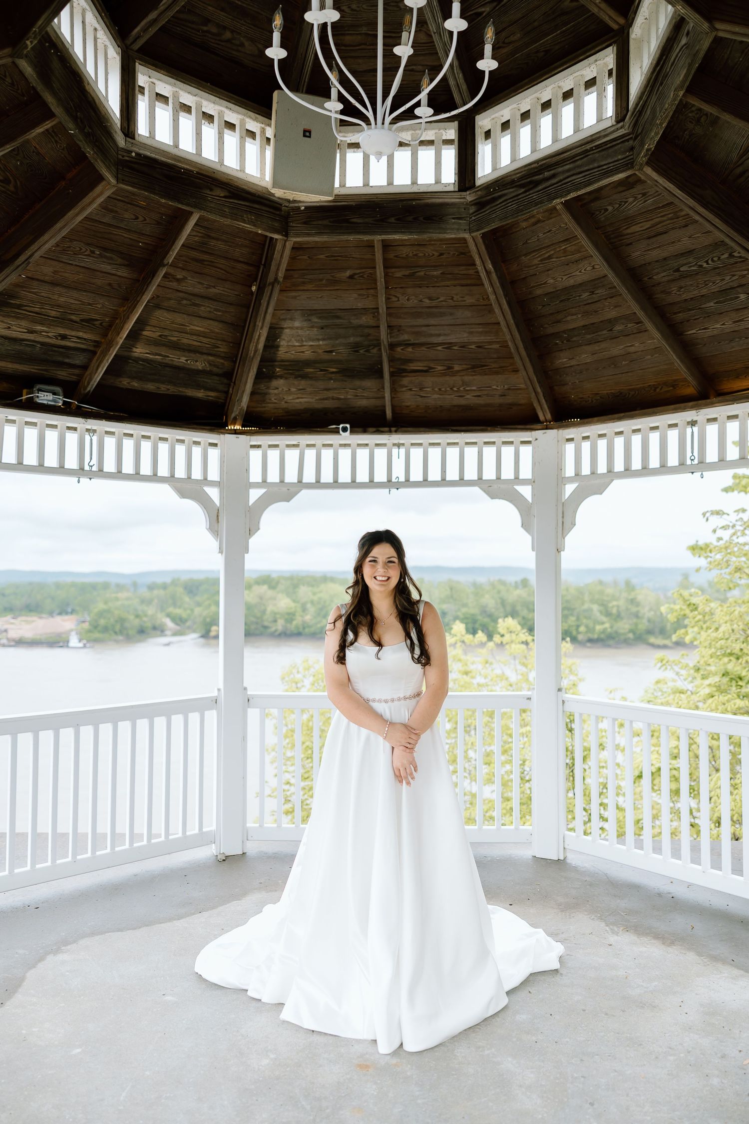 A bride in a flowing white wedding dress poses in an elegant white gazebo with panoramic views of nature.