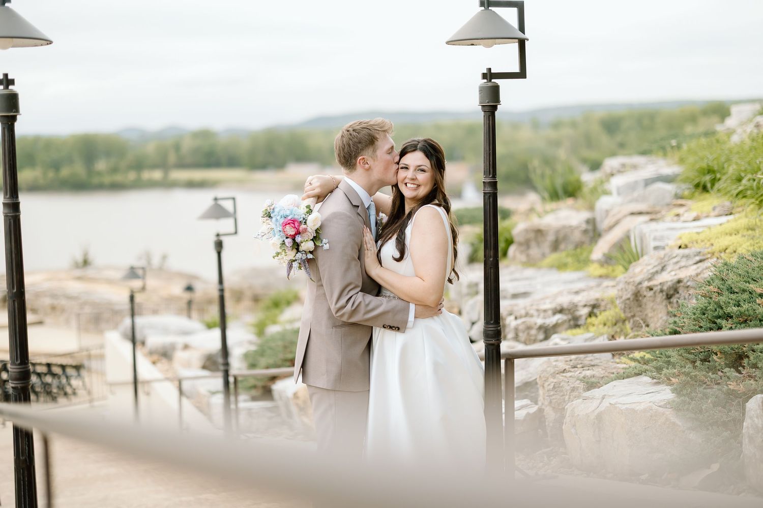 A romantic wedding couple embraces on a scenic overlook with lake views and lamp posts in the background.