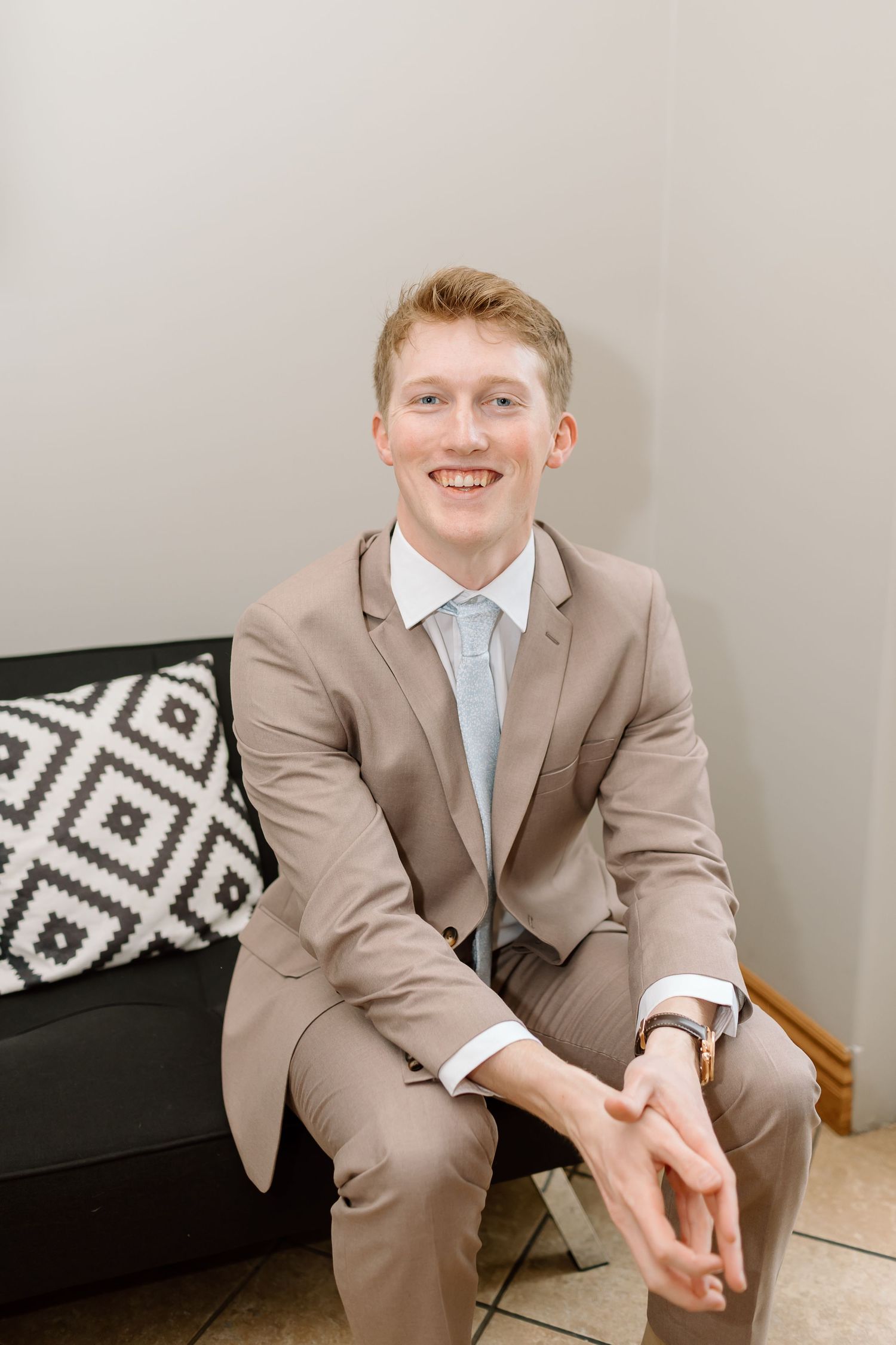 A person in a beige suit and light blue tie sits on a couch with a black and white geometric patterned pillow.