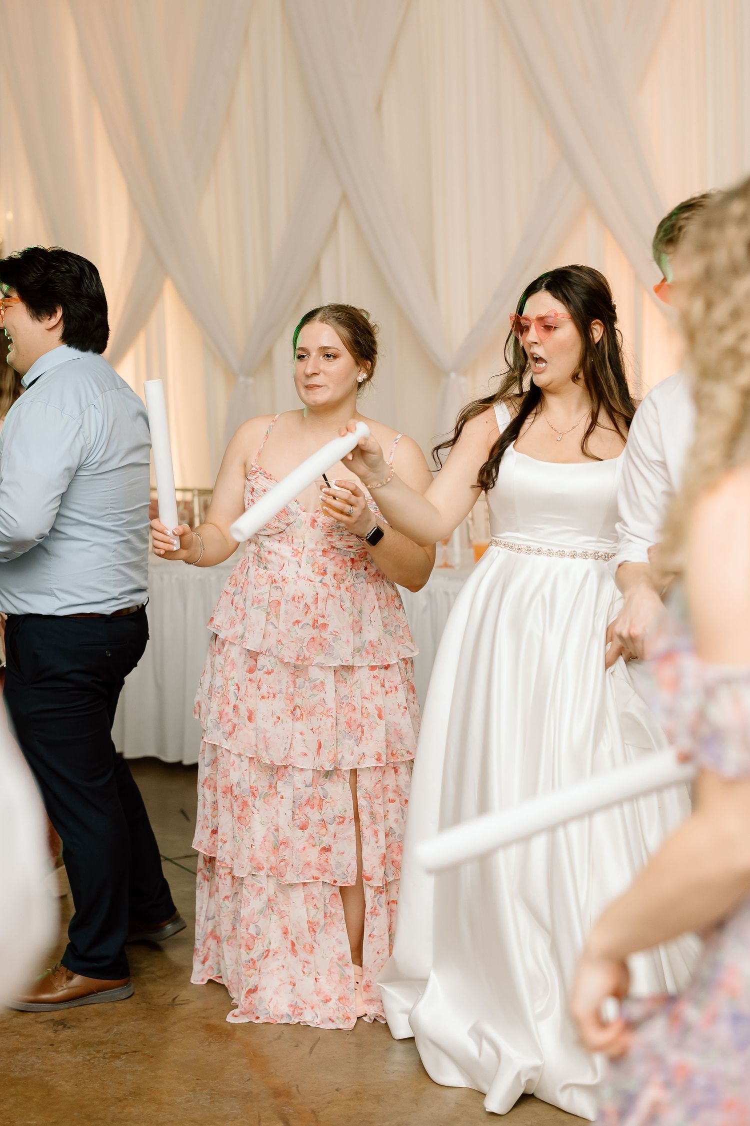 Wedding guests dance together at a reception with white drapery in the background and floral pink dresses.