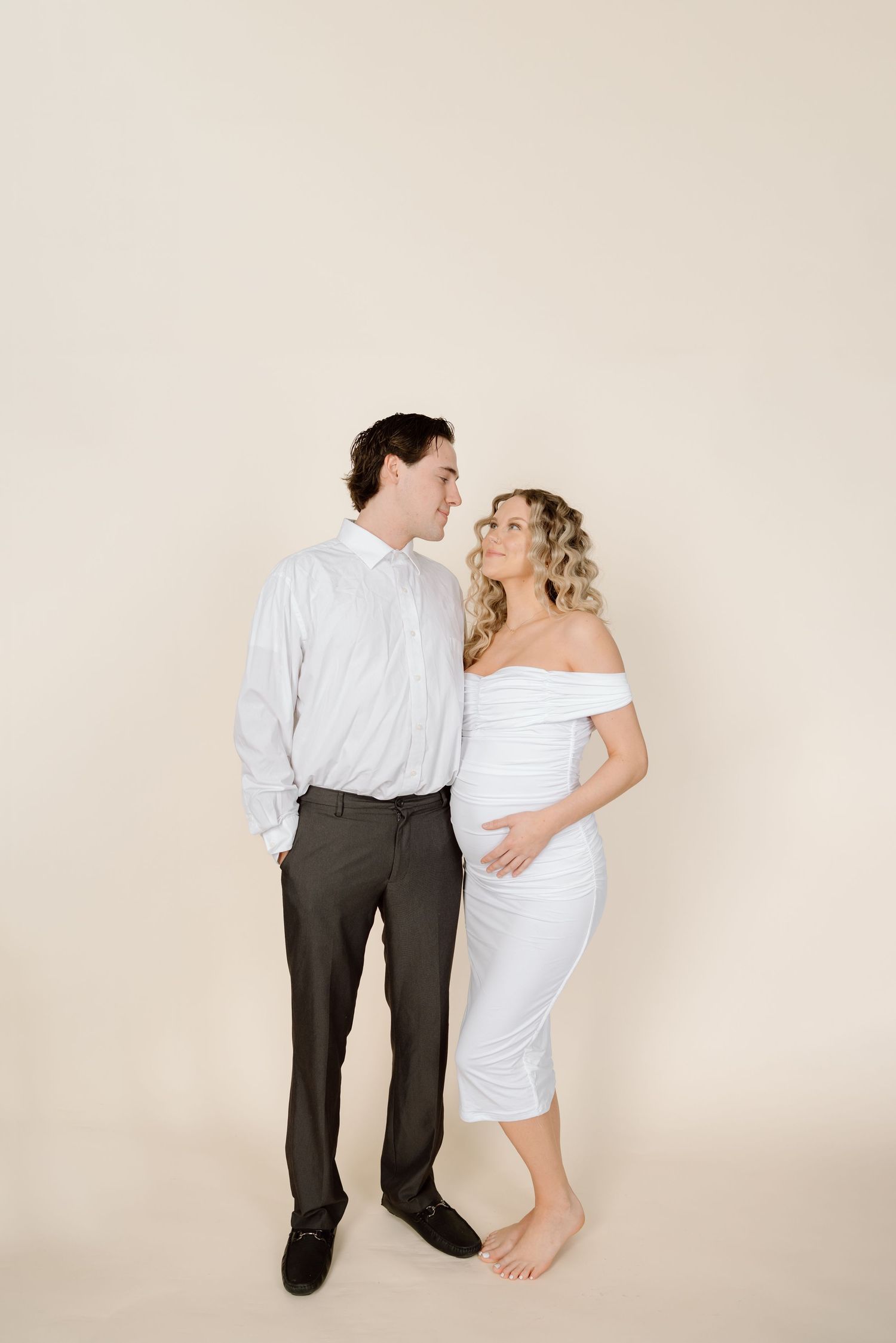 A couple shares an intimate moment against a beige backdrop, with the woman wearing white and the man in business casual attire.