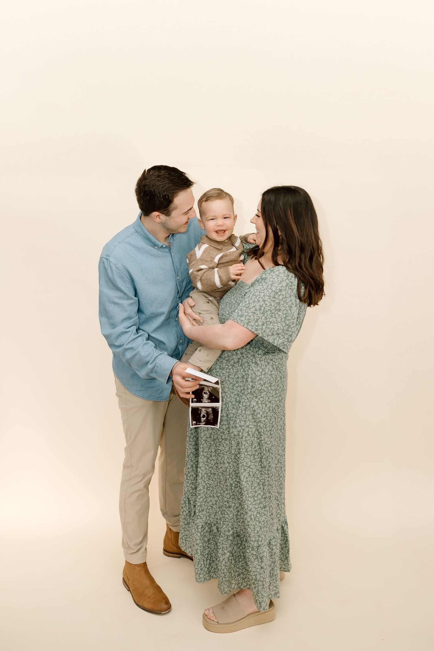 Family portrait with couple in casual attire holding baby and ultrasound photo against neutral background.