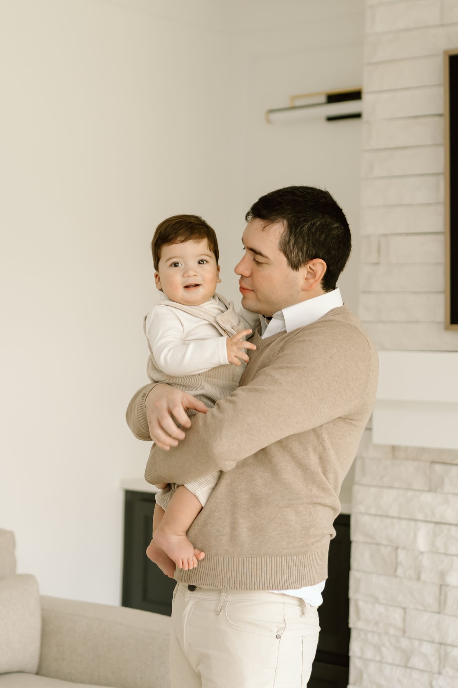 A parent in a beige sweater tenderly holds a baby wearing white against a light-colored brick wall interior.